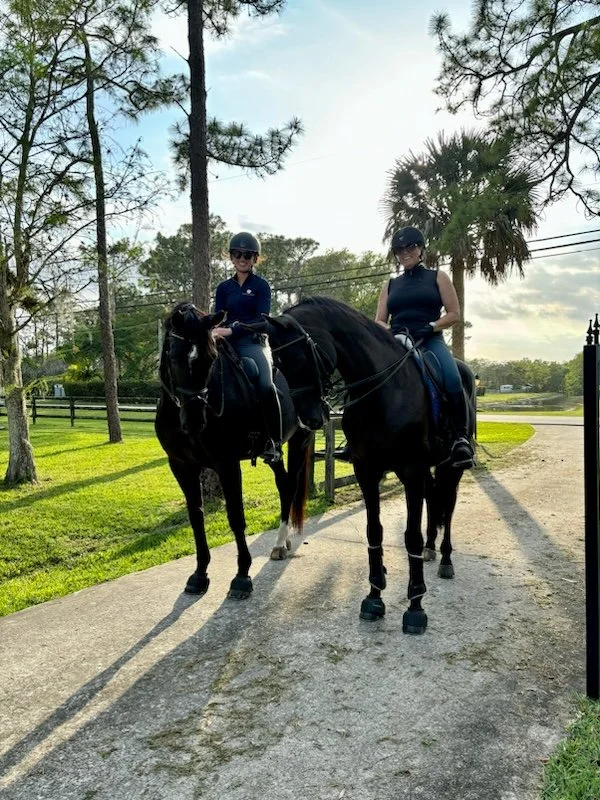 Two women wearing helmets and riding Western-style saddles on black horses on a dirt trail, with trees and a blue sky in the background.
