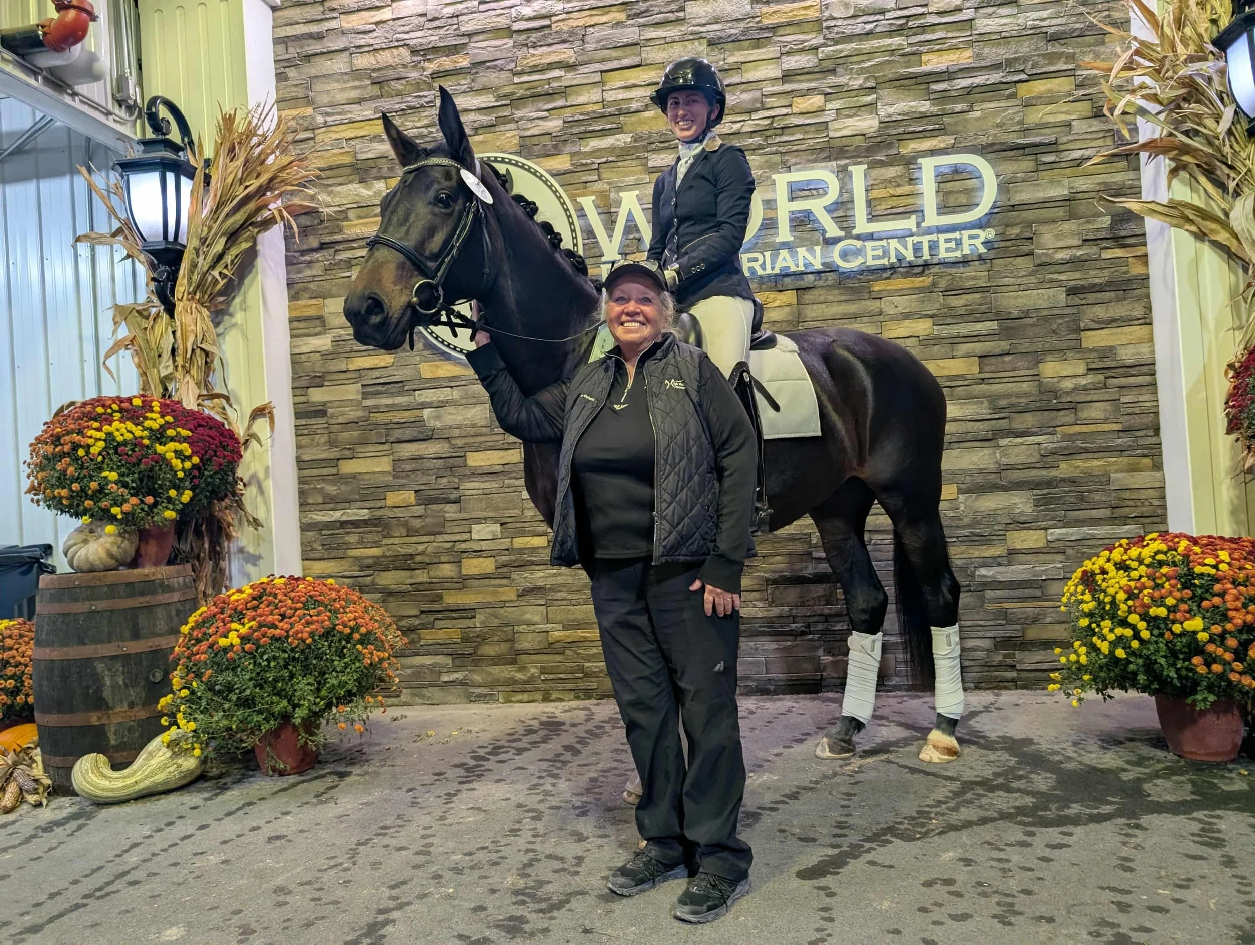 A woman standing in front of a rock wall, holding a smiling rider on a horse. The rider wears a helmet and riding attire. The horse is dark-colored with white leg wraps. The scene is decorated with autumnal plants and flowers, and a sign behind reads