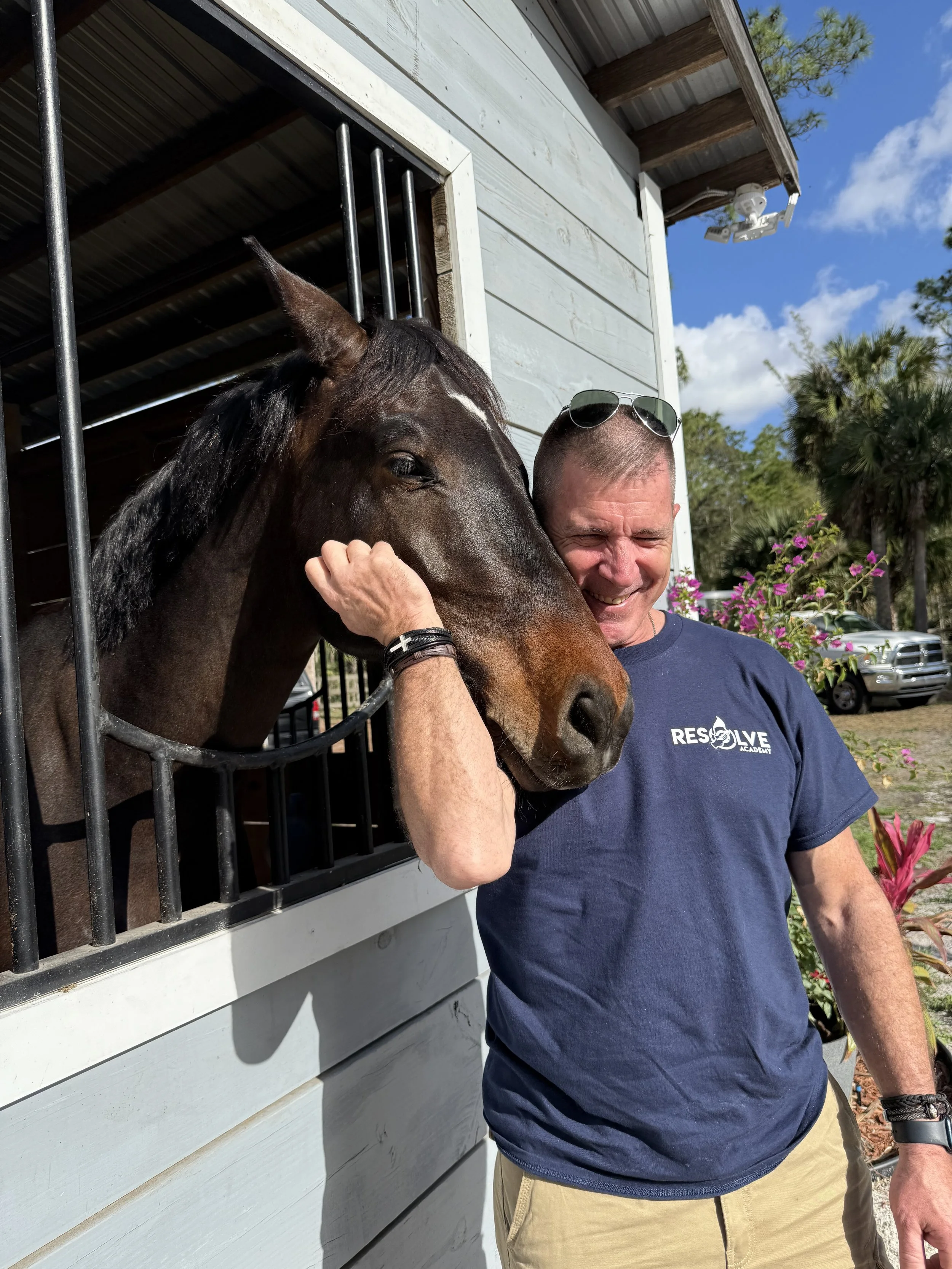 A man with sunglasses on his head hugging a horse outside a stable on a sunny day, with trees and flowers in the background.