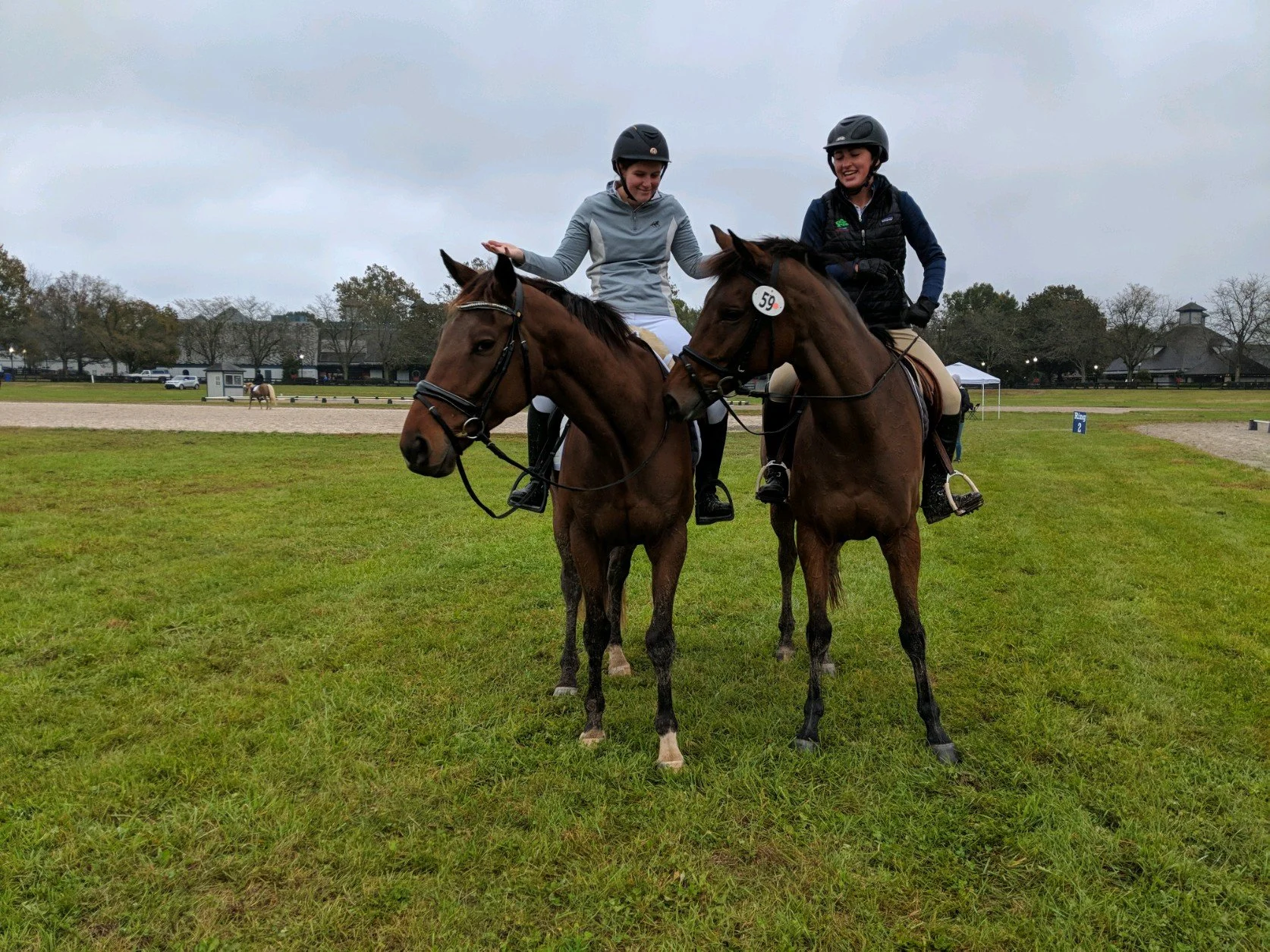 Two women riding horses on a grassy field, one adjusting her saddle while the other laughs, both wearing helmets and riding gear, overcast sky in the background.
