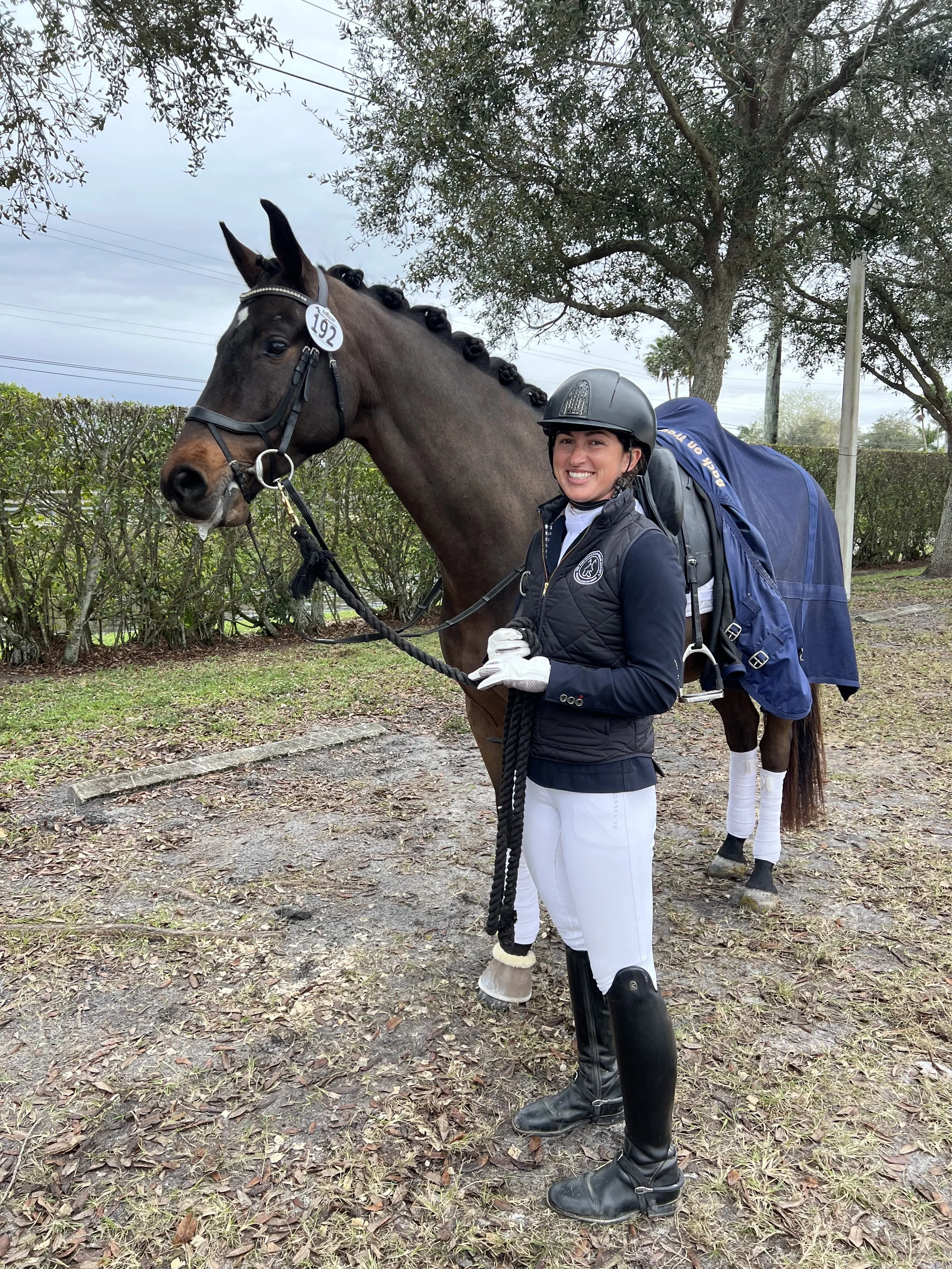 A woman dressed in equestrian attire, including a helmet, riding boots, and white breeches, stands smiling with a dark brown horse with braided mane. The horse has a blue blanket draped over its back and white leg wraps.