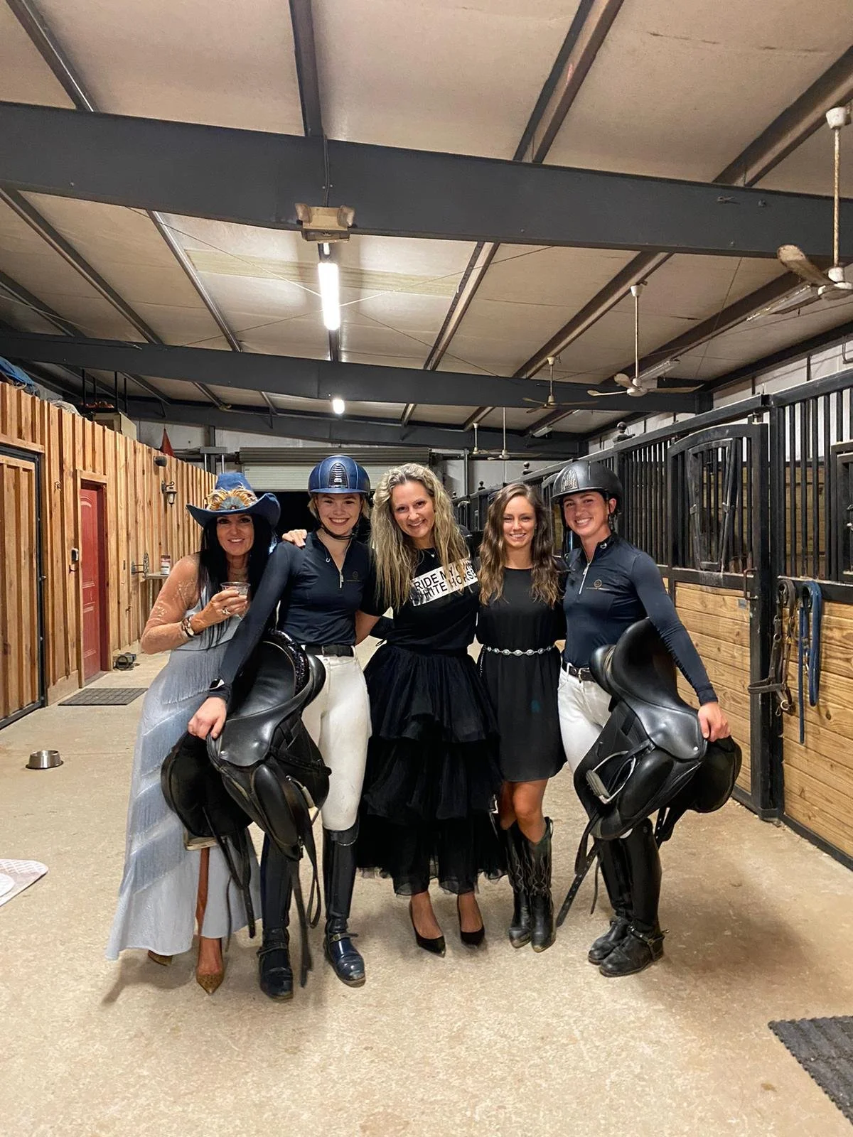 Group of five women in riding attire and equestrian gear, standing together in a stable, smiling at the camera.