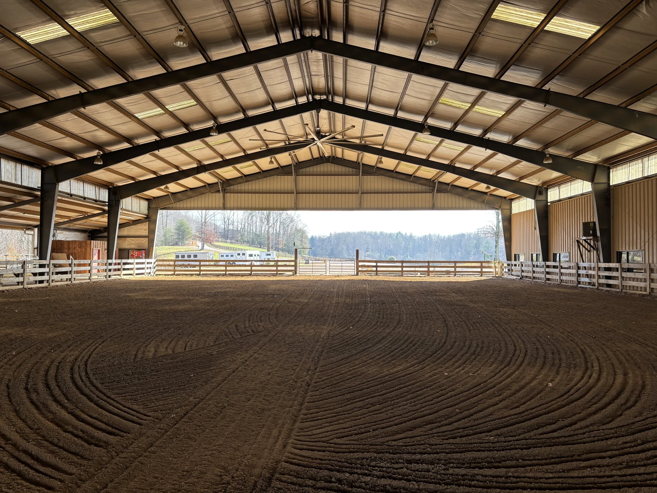 Interior of an indoor riding arena with freshly groomed dirt floor and wooden railings, open at one end revealing outdoor scenery with trees and a horse trailer.