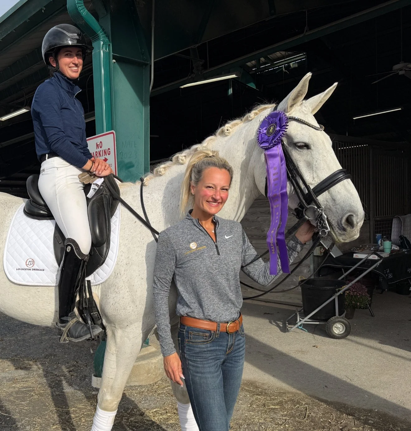 A woman in athletic wear standing beside a white horse with a purple ribbon and rosette on its bridle, while another woman sits on the horse in riding attire and helmet.