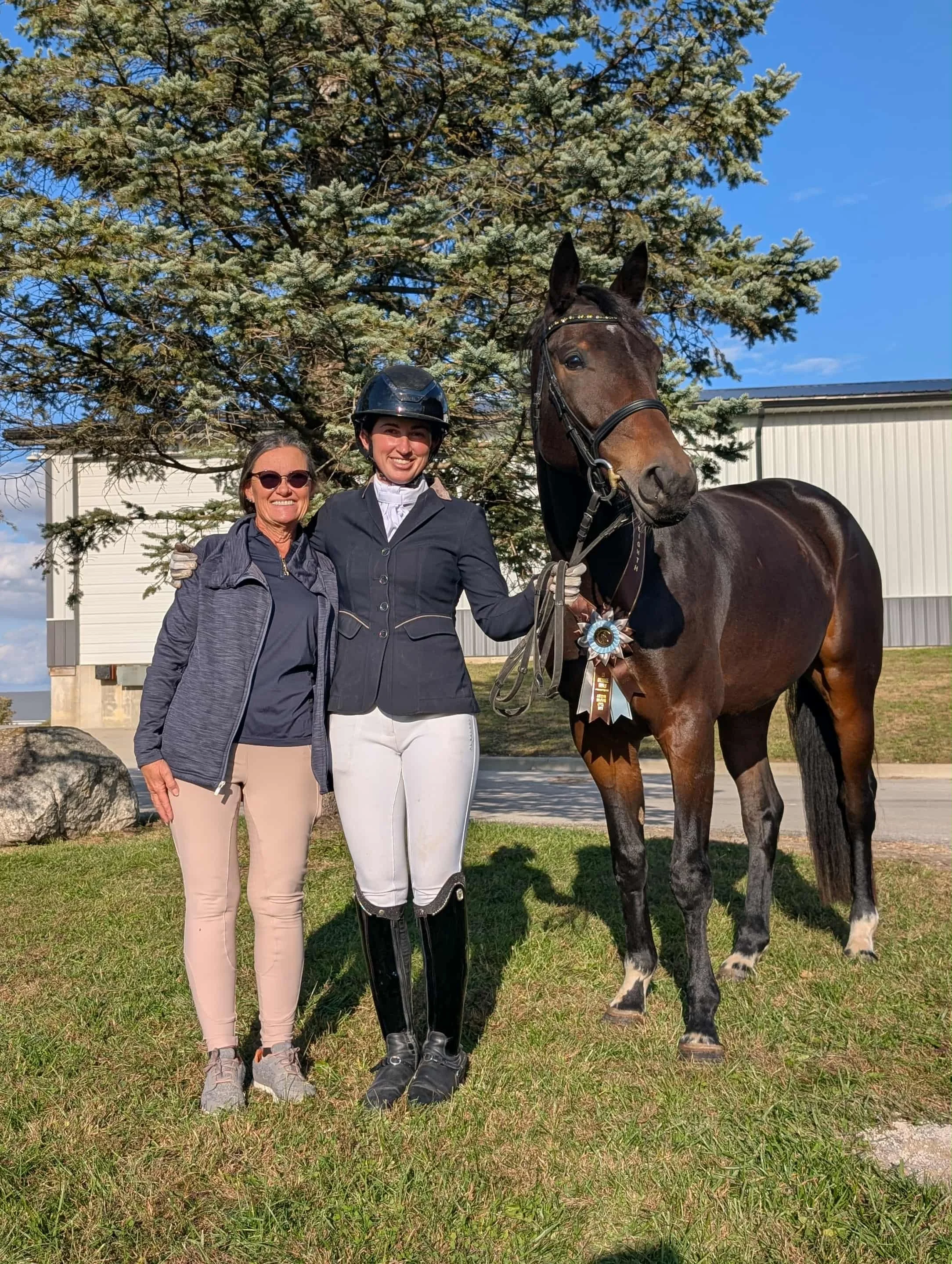 Two women and a horse standing together outdoors near a tree, with a building in the background. The woman on the right is in riding attire, holding the horse's reins, while the woman on the left is dressed casually. The horse has a ribbon attached t