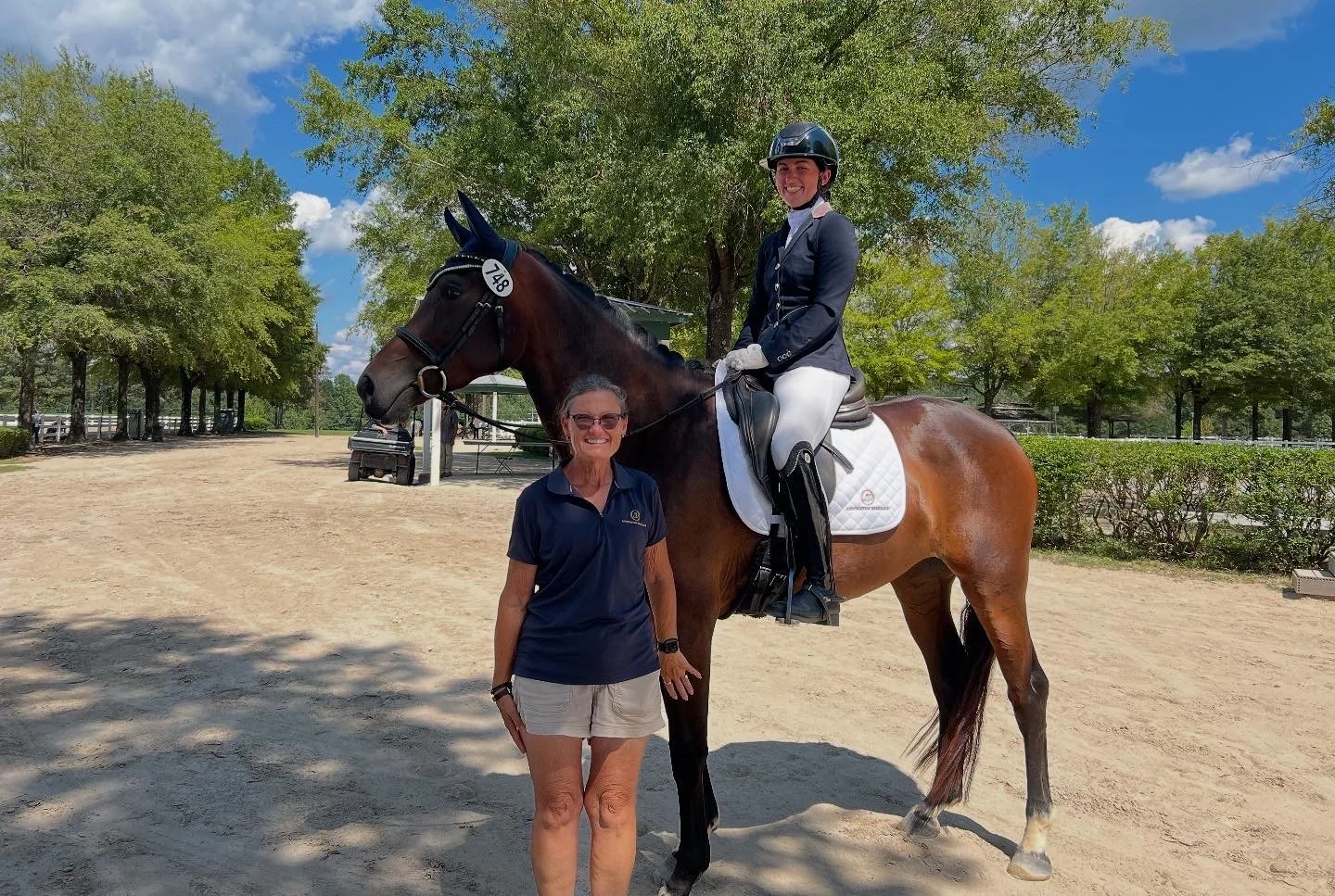 A woman standing next to a person on a horse in an outdoor riding arena with trees and blue sky in the background.