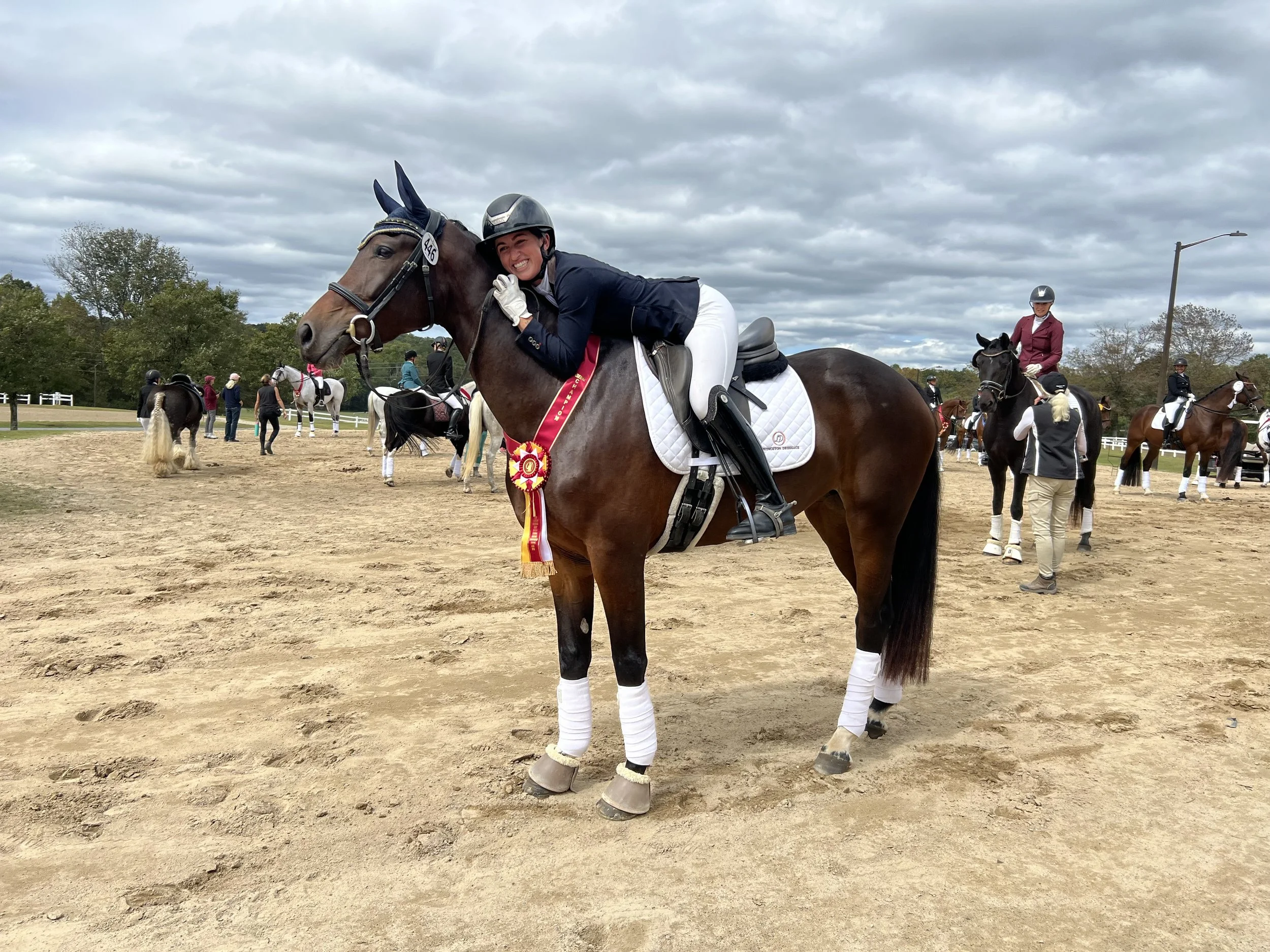 A female equestrian rider in formal riding attire cuddles her brown horse with a white blaze on its face after a competition, wearing a ribbon and rosette. Other riders and horses are visible in the background on a sandy outdoor arena under a cloudy 