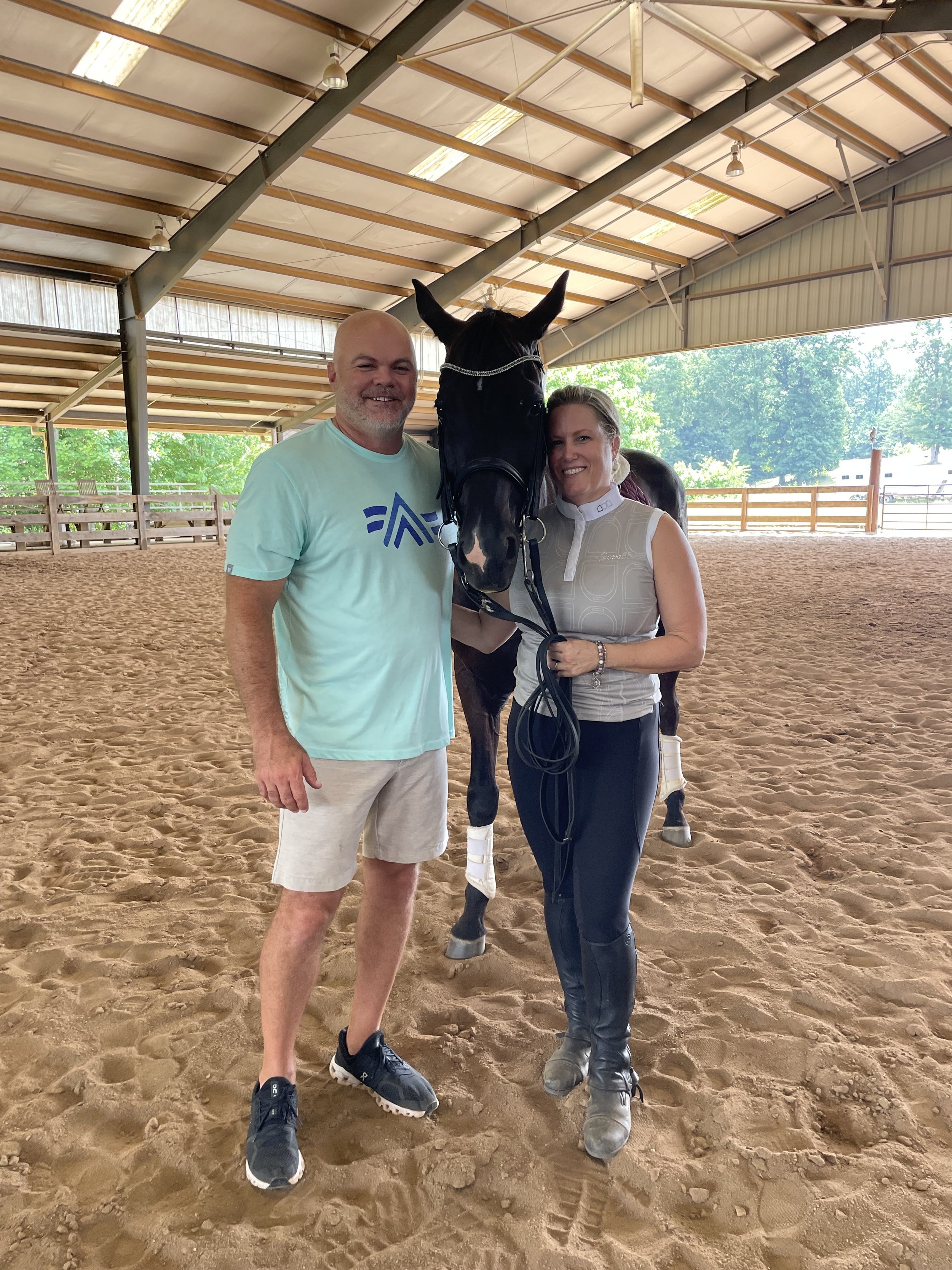 Two people and a horse in a covered indoor arena. The man is wearing a light blue shirt and shorts, and the woman is in riding attire. They are smiling and standing close with the horse between them, which has a bridle and leg wraps.