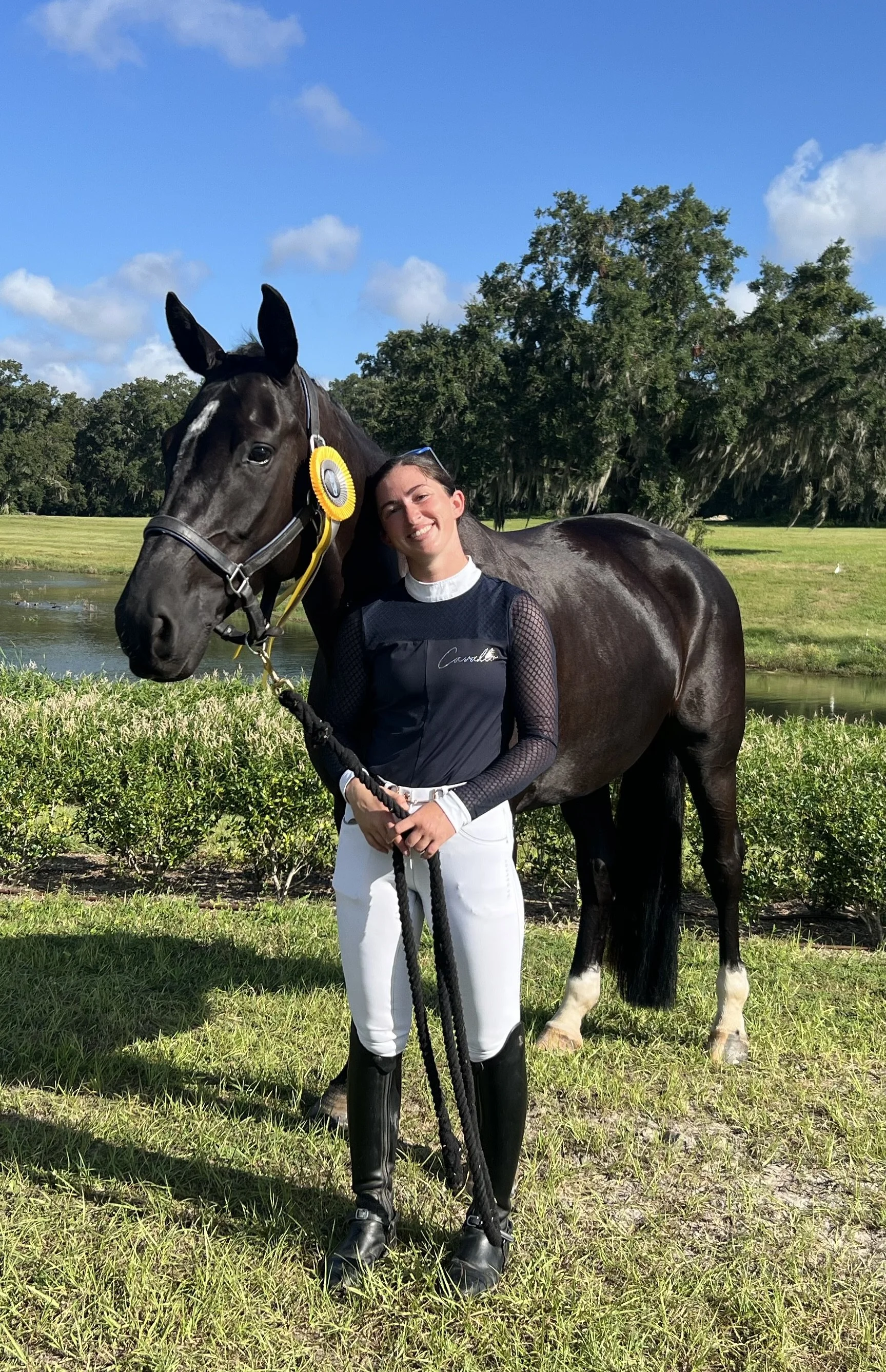 A young woman in equestrian attire stands with a black horse holding the horse's reins, both outdoors near a pond, trees, and a grassy field, under a blue sky with clouds.