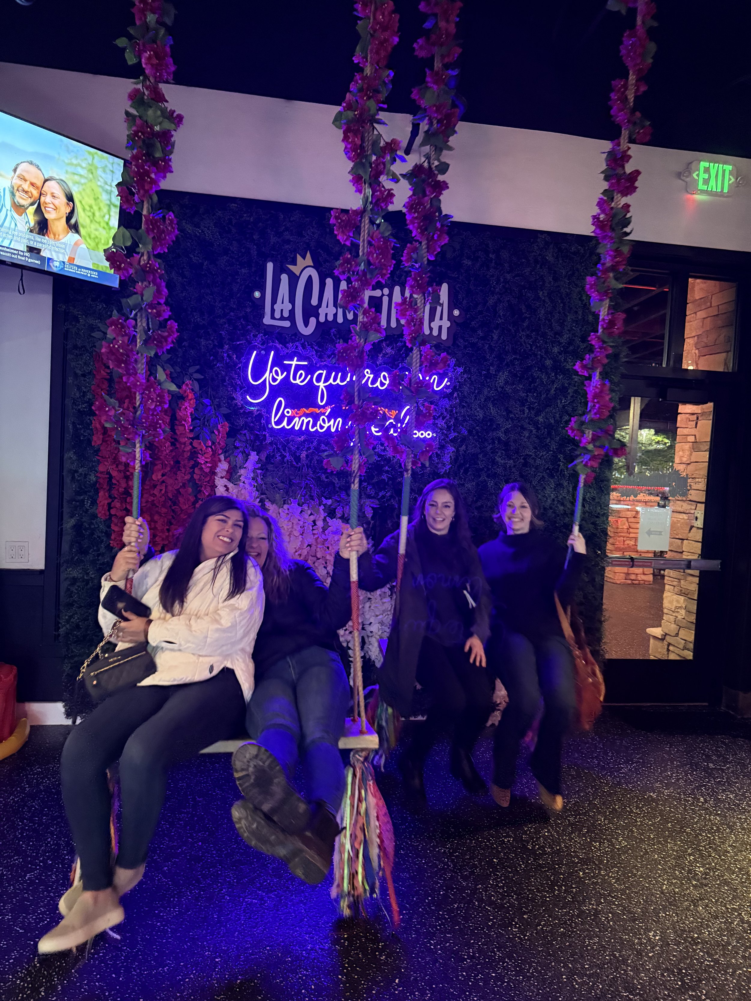 Four women sitting on decorative swings with pink flowers, inside a restaurant with a dark wall and neon sign, celebrating together.