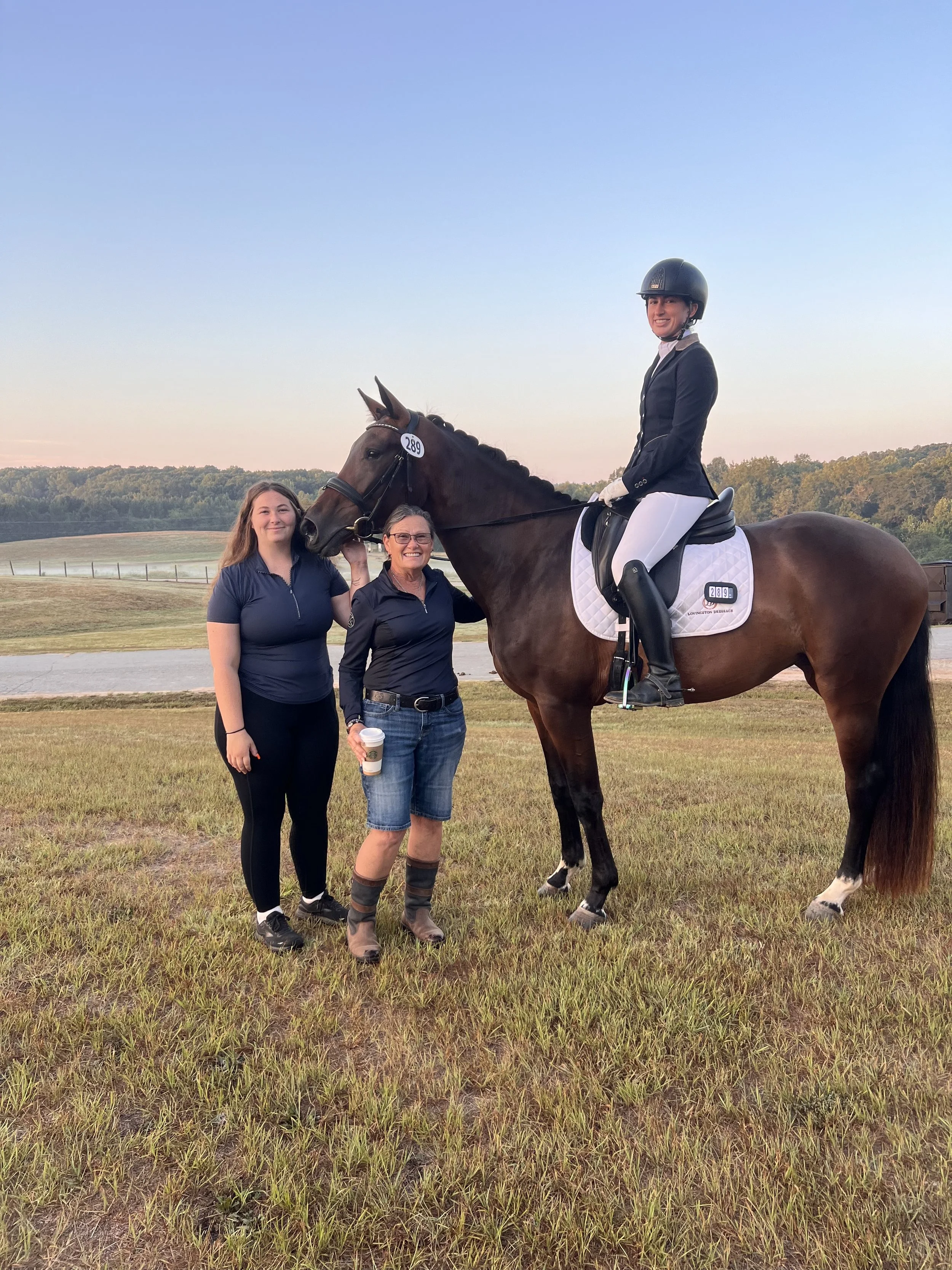 Three women and a rider on a horse outdoors at sunset, with a grassy field and trees in the background.