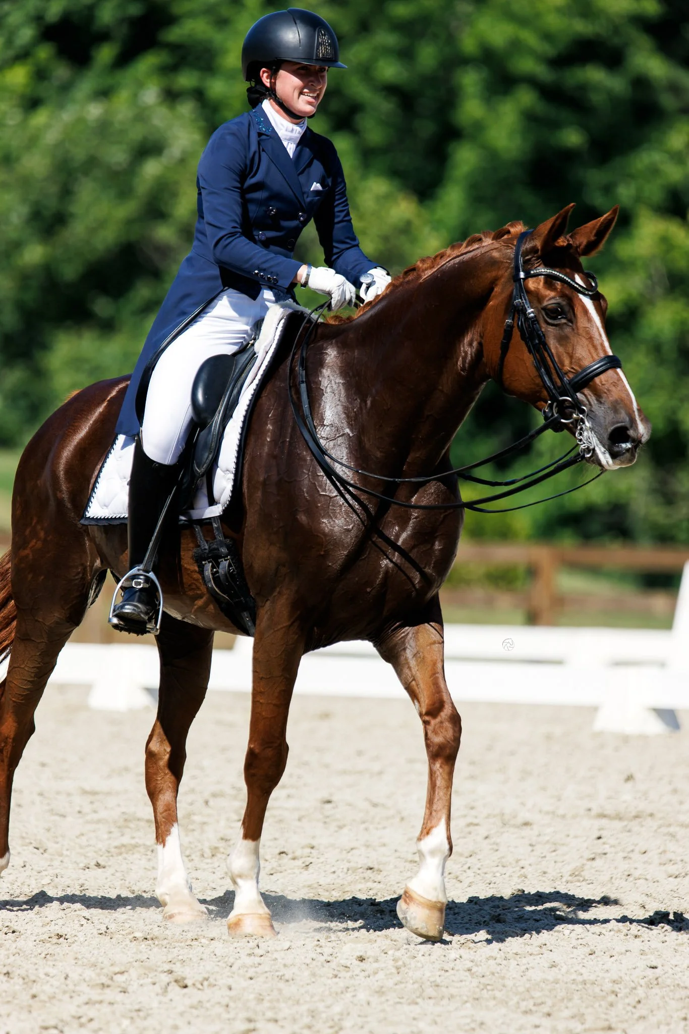 A woman in riding gear, including a helmet and riding jacket, smiling while riding a brown horse with a white marking on its face during a dressage event.
