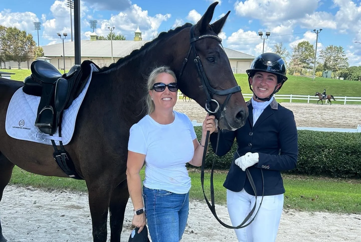 Two women standing with a dark brown horse at an equestrian facility on a sunny day; one woman is in casual attire with sunglasses, the other is in riding equestrian outfit with a helmet, both smiling.
