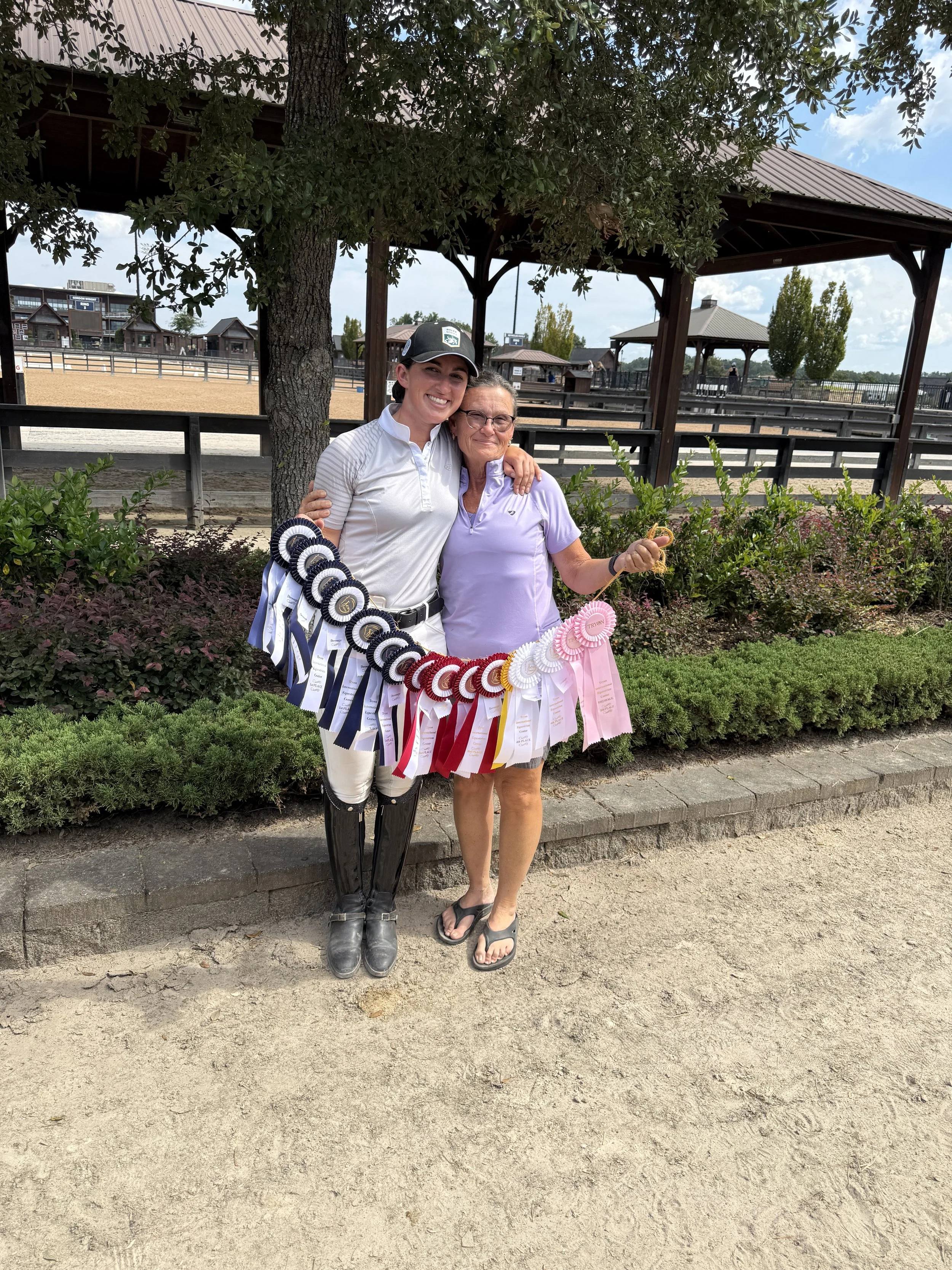 Two women standing outdoors near a horse riding arena, smiling and holding a large display of blue, red, and pink ribbons.