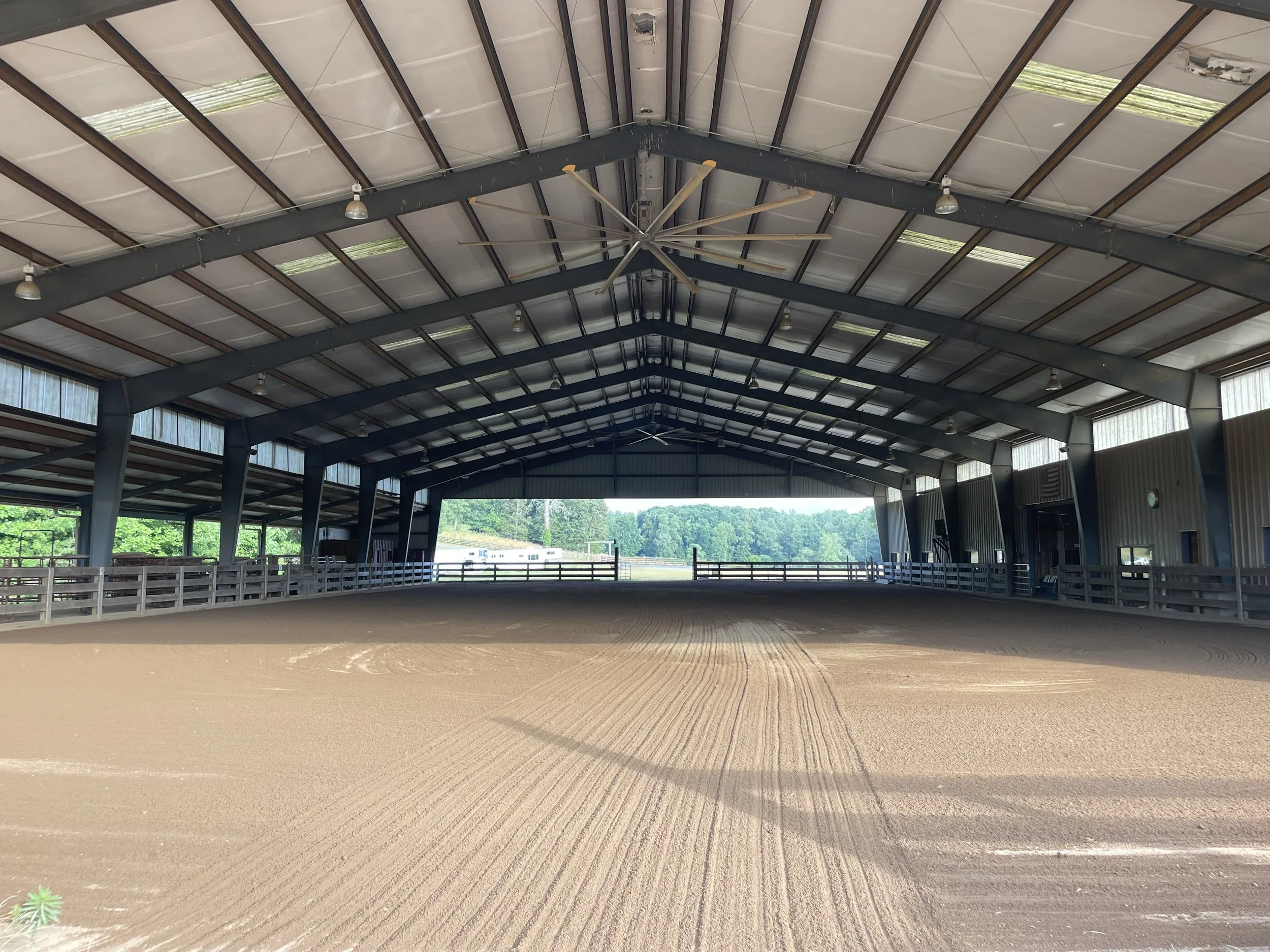 Sand riding arena inside a large covered structure with open sides and metal roof, surrounded by green trees.