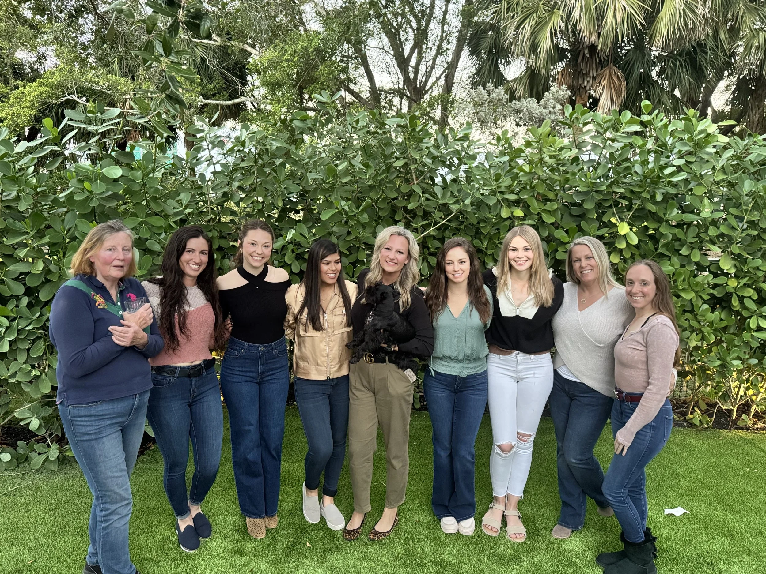 A group of nine women standing outdoors on grass in front of green bushes and trees, smiling at the camera.