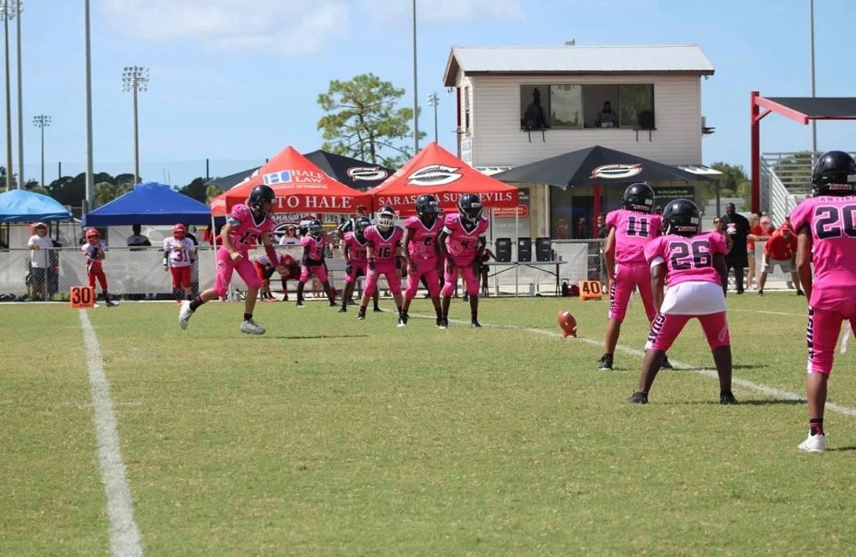Football players in pink uniforms line up on the field during a game, with a scoreboard, tents, and spectators in the background on a sunny day.