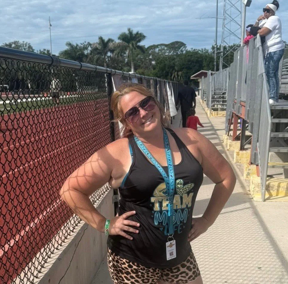 Smiling woman with sunglasses standing at a sports stadium or event, wearing a tank top, patterned shorts, and a lanyard around her neck.