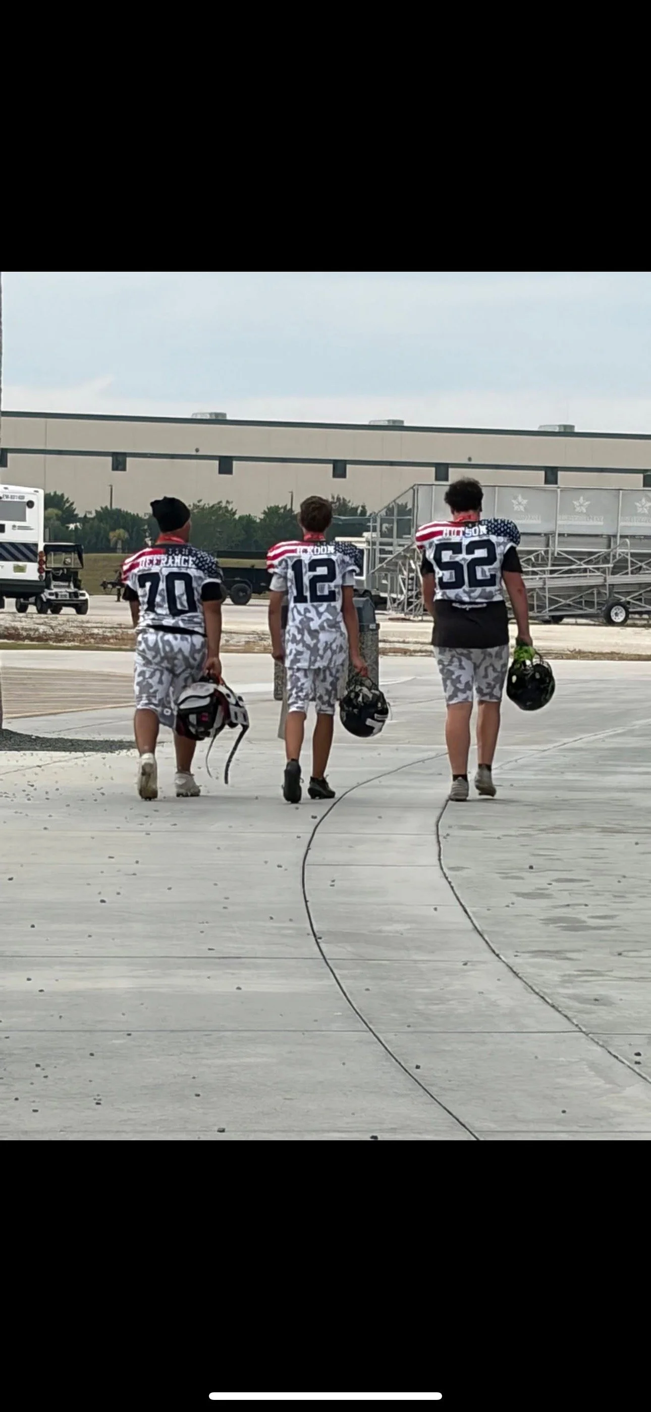 Three young football players walking away from the camera, carrying helmets, wearing matching jerseys with the American flag design, numbered 70, 12, and 52, on a tarmac at an airport.