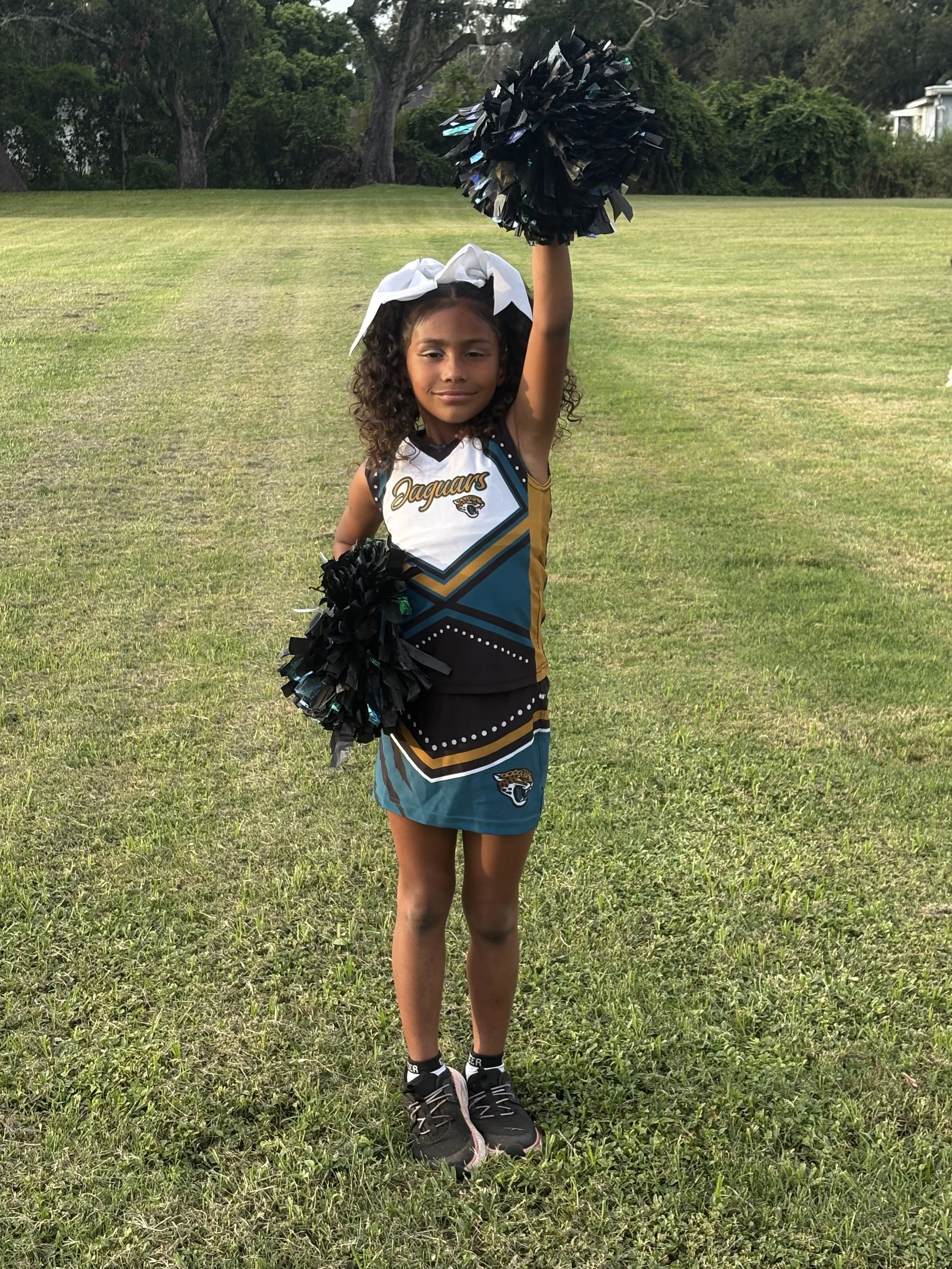 Young girl in a Jaguars cheerleading outfit holding black pom-poms, standing on a grassy field.