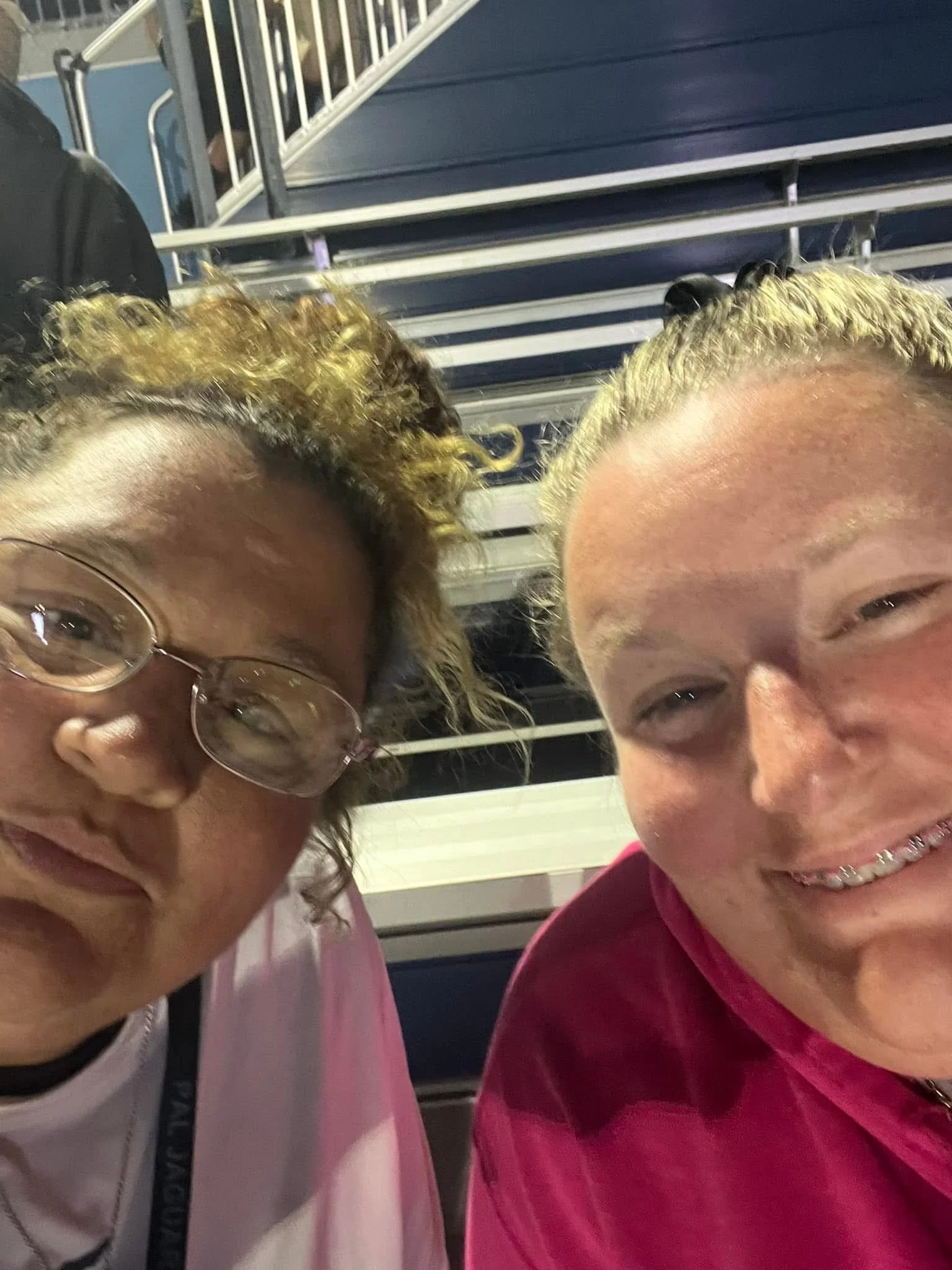Two women taking a close-up selfie, sitting in stadium seats with metal railings in the background. One woman has curly hair and glasses, the other has blonde hair tied back, both smiling.