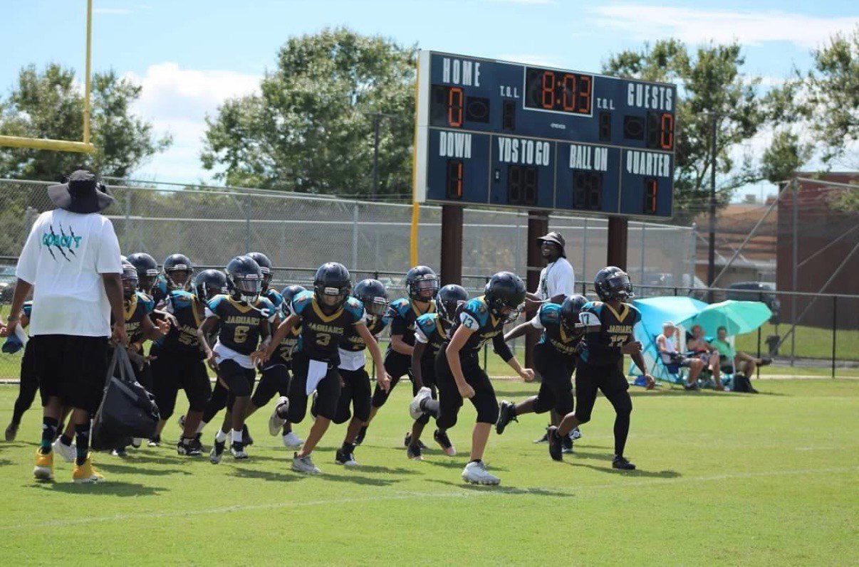 Youth football team in black jerseys and helmets running onto the field for a game, coaches and spectators under a bright blue sky.