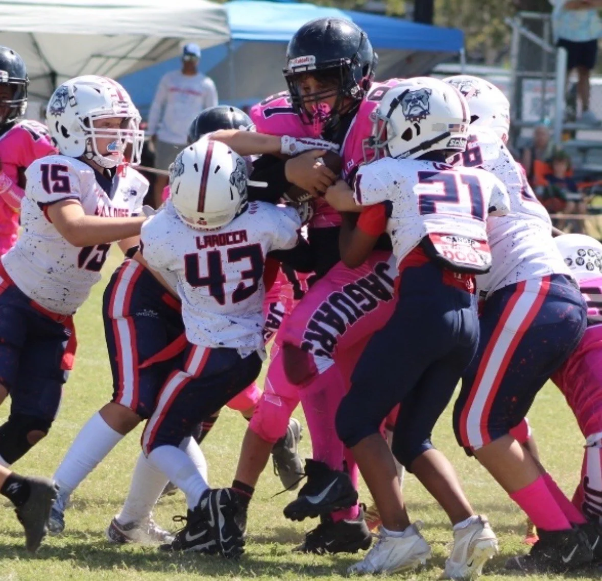 Youth football players in white and pink uniforms tackle a player in a black and pink uniform during a game.