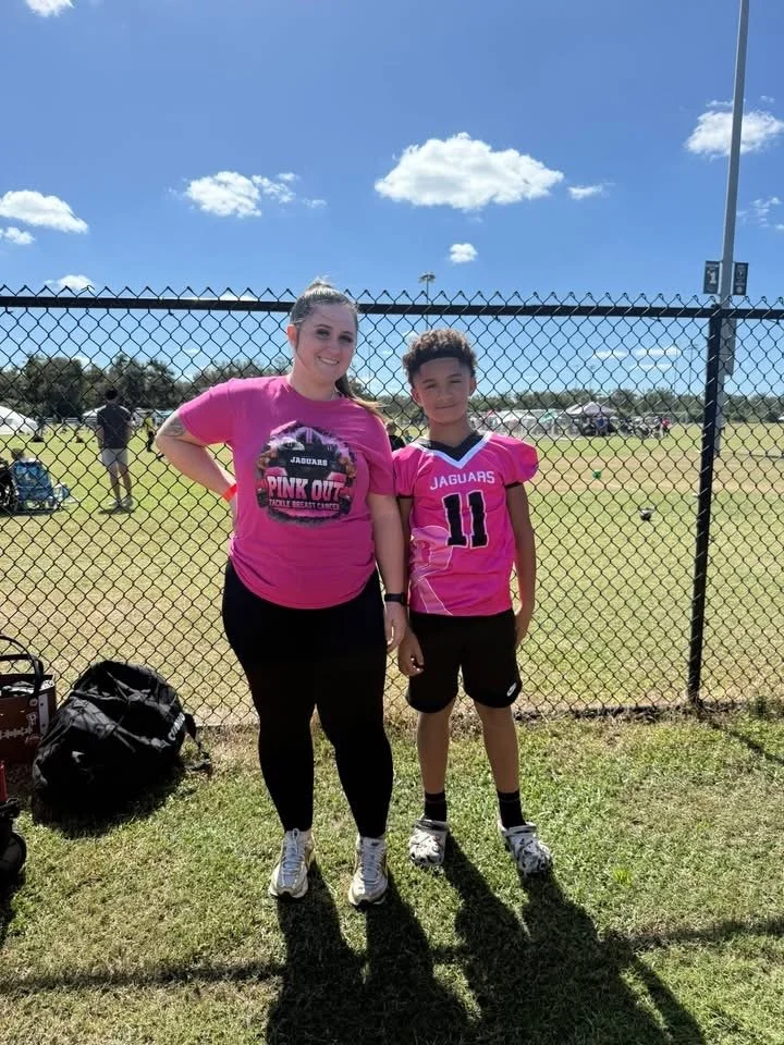 A woman and a young boy standing in front of a chain-link fence at a sports field, both wearing pink jerseys; the woman’s shirt has a Jaguars logo and the words "Pink Out," and the boy’s shirt has "Jaguars" and the number 11.