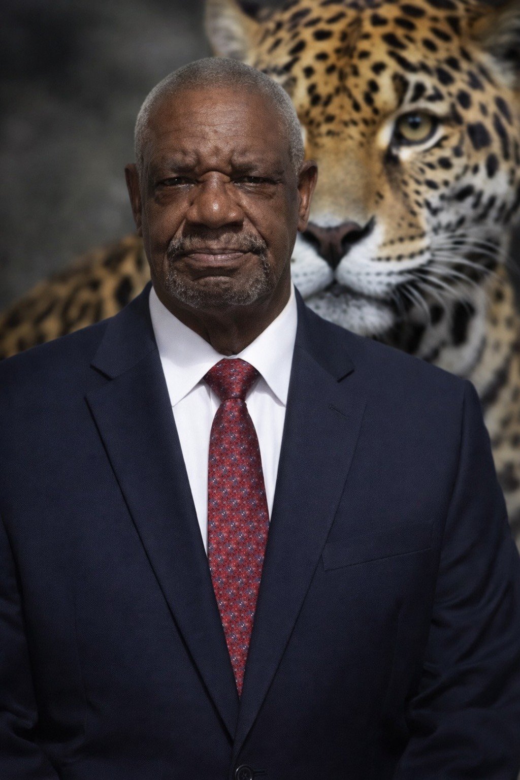 man in a dark suit and red tie stands in front of a large photograph of a jaguar
