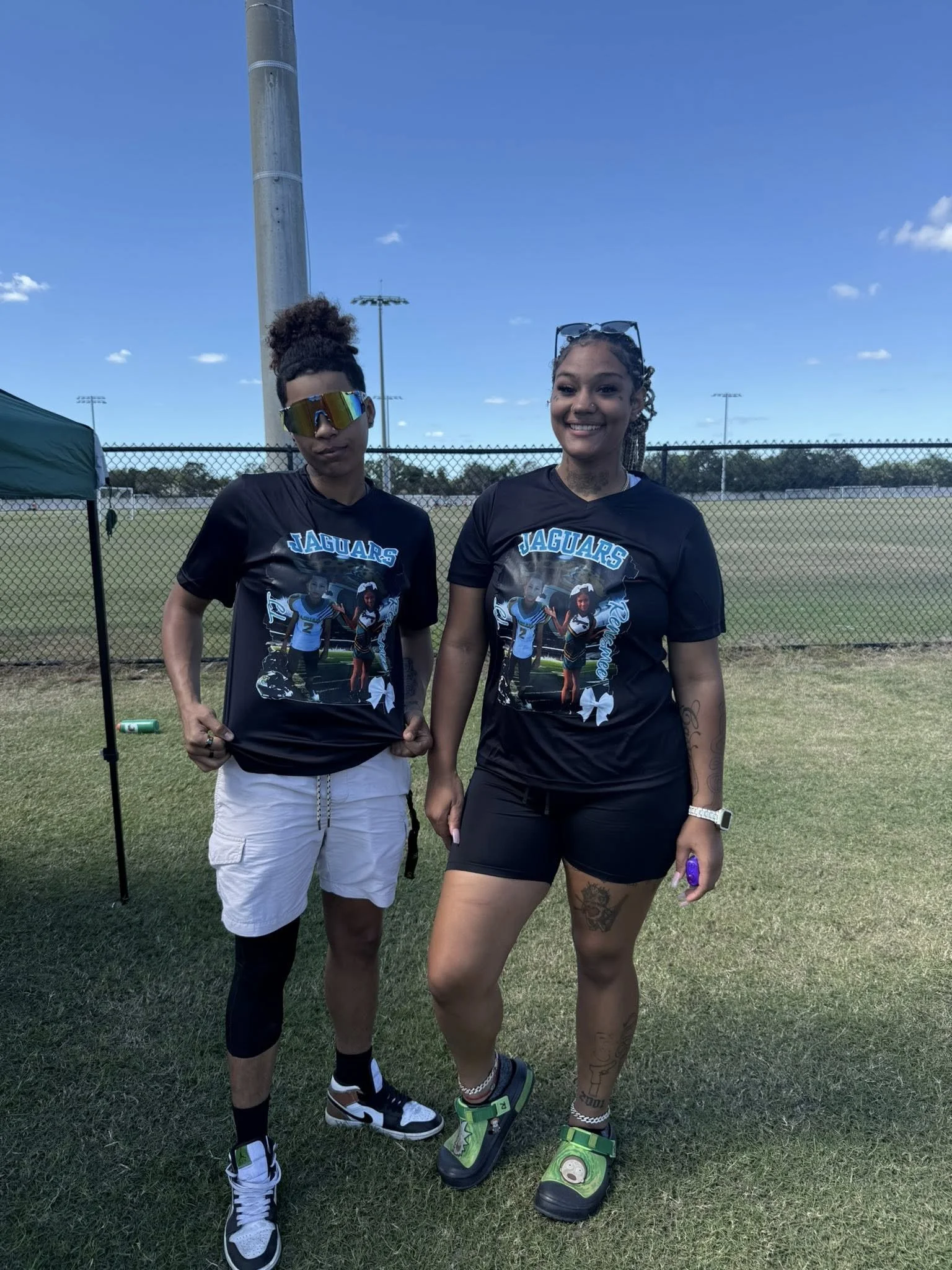 Two women standing on a football field, wearing matching 'Jaguars' t-shirts with a photo and the number 2 on them. One is wearing white shorts, black leggings, and colorful sneakers; the other is wearing black shorts, sneakers, and has tattoos on her