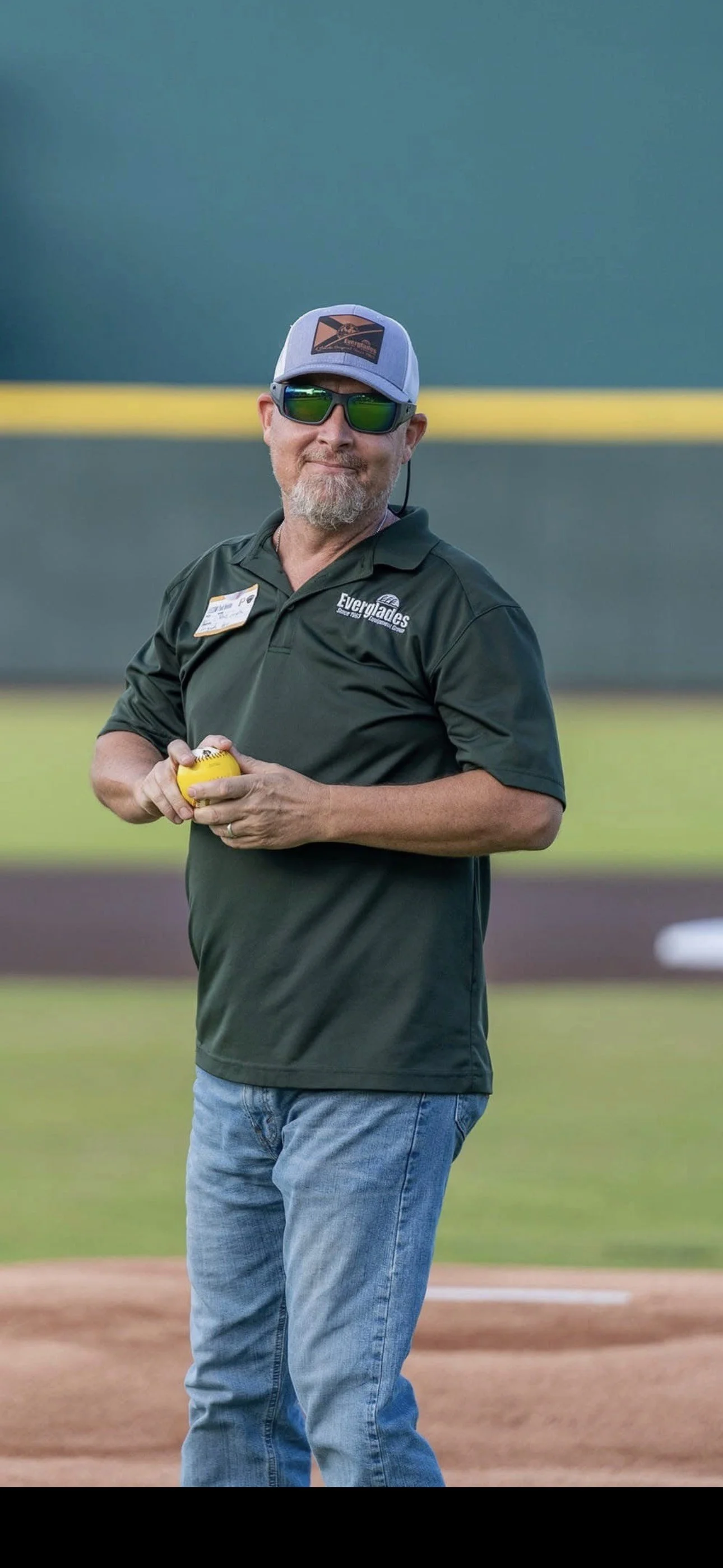 A man standing on a baseball field, wearing sunglasses, a cap, a dark green polo shirt with a logo and name tag, and holding a yellow baseball.