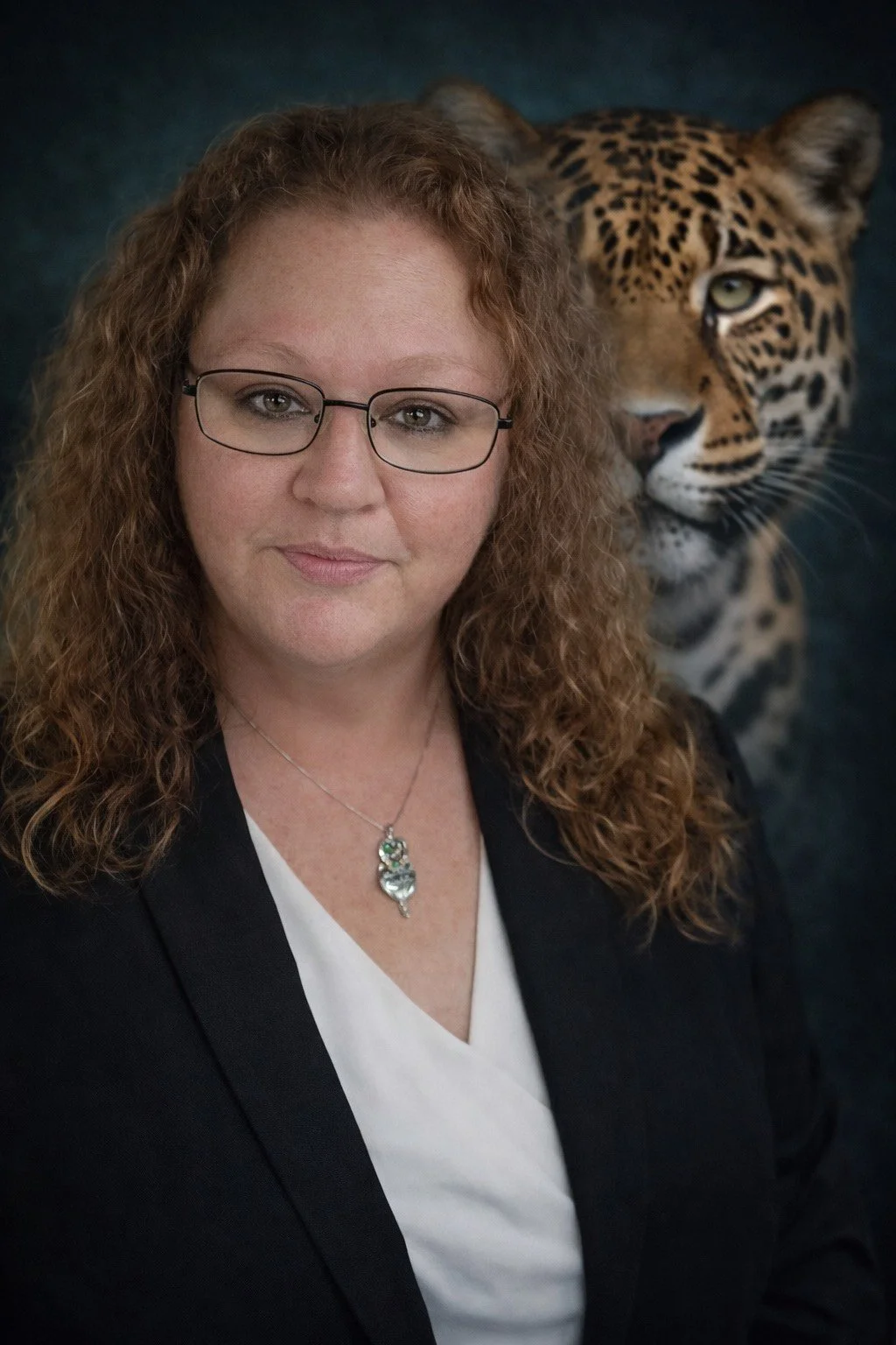 A woman with curly red hair, glasses, and a black blazer, wearing a necklace, standing in front of a backdrop with a close-up of a leopard's face.