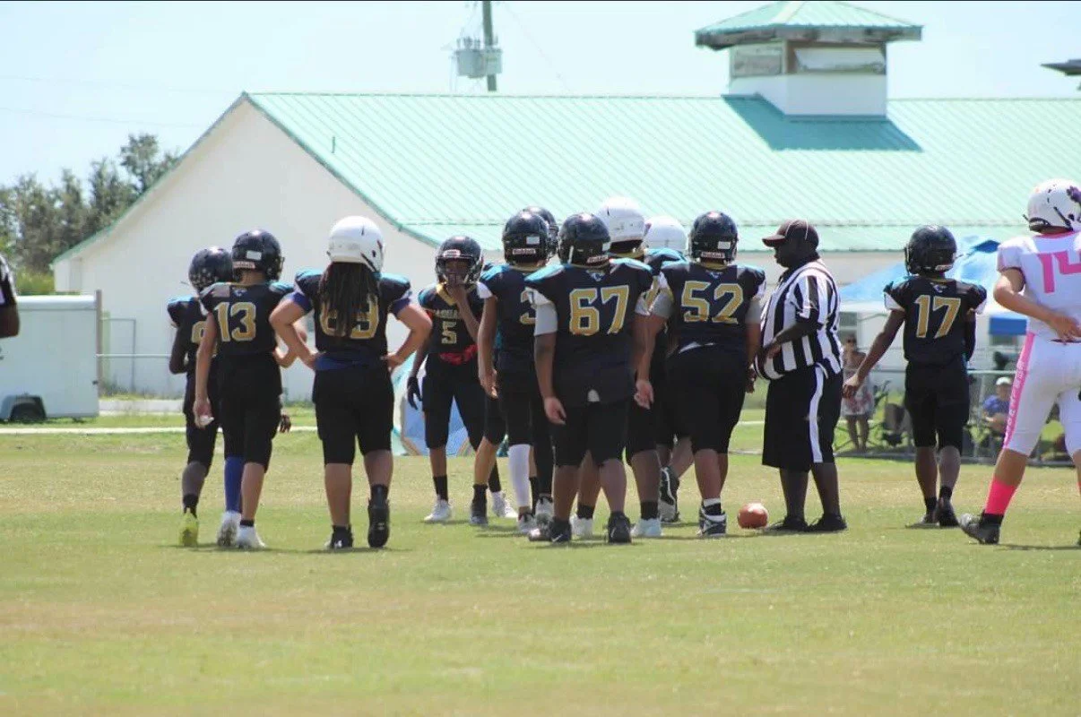 Youth football team huddled on the field with a referee nearby during a game. Manatee Jaguars football 