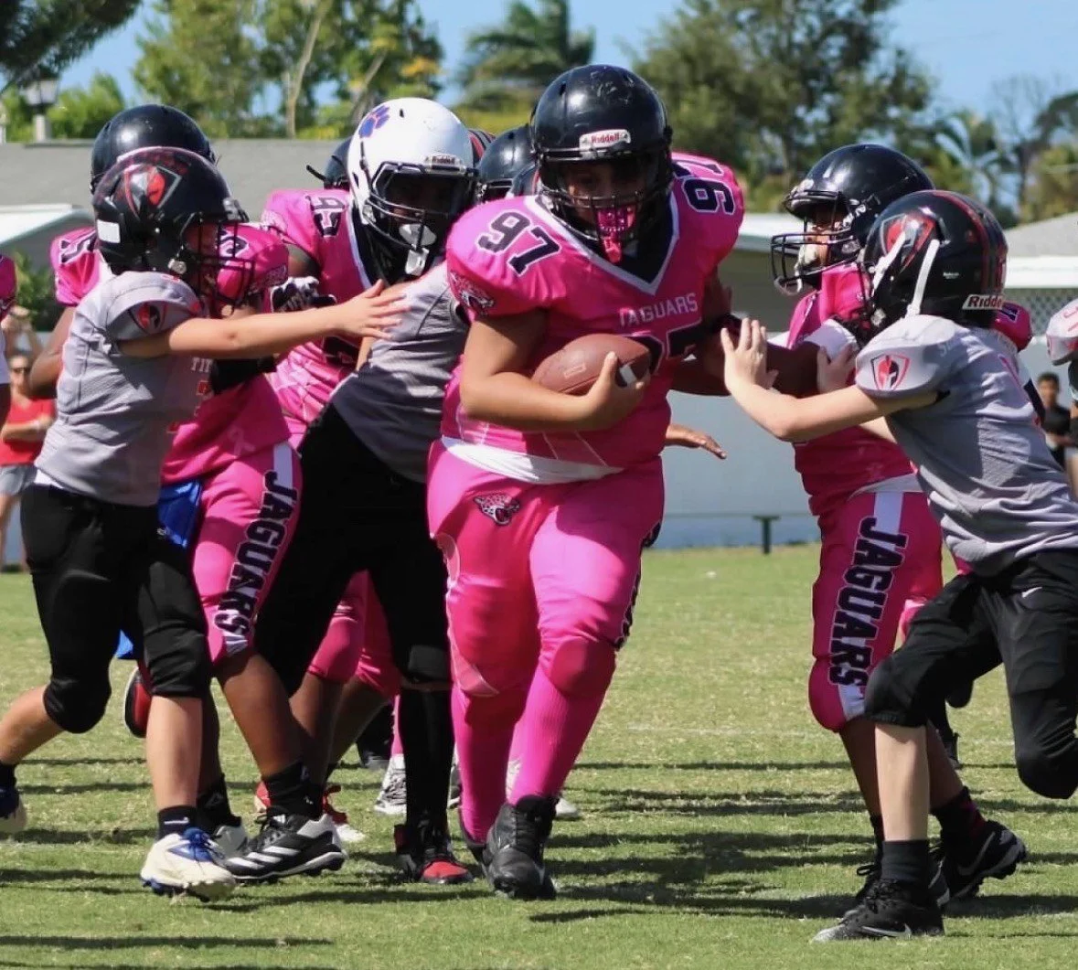 Youth football game with players in pink and gray jerseys, helmets, and pads on a grassy field.