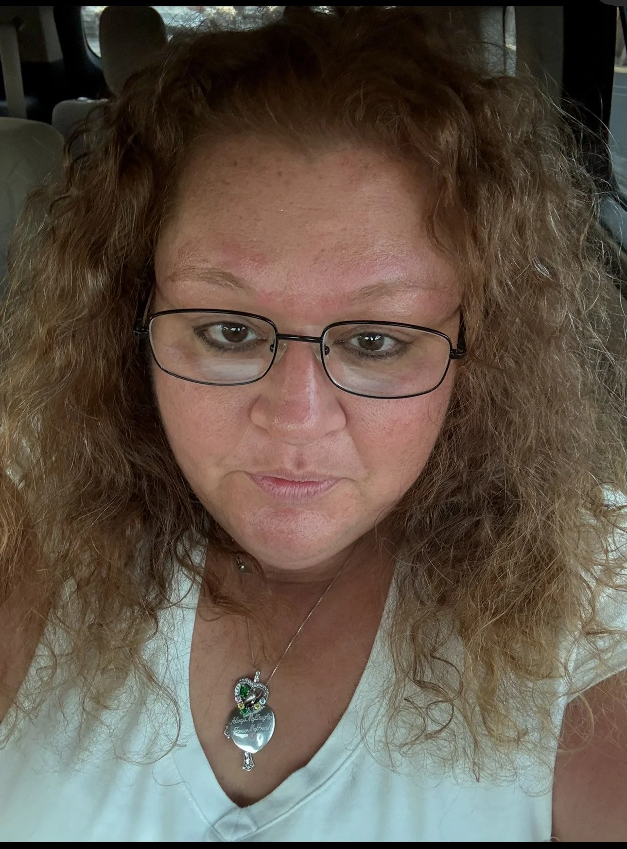 Close-up of a woman with curly, reddish hair, glasses, and a necklace, sitting inside a vehicle.