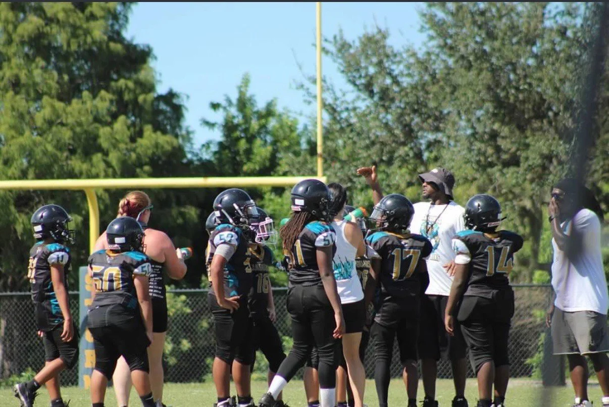 Youth football team in black uniforms with helmets, gathered on field during practice or game with coaches.