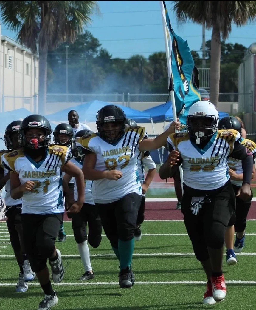 Youth football team running onto the field with a flag at a football game.