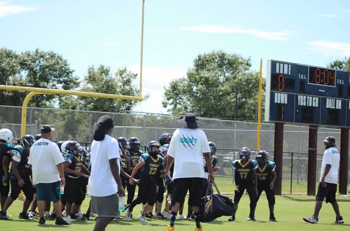 Youth football team practicing on a field during daytime, coaches present, scoreboard showing 8:24, blue sky with some clouds.