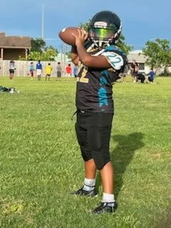 A young football player standing on a football field, holding a football in both hands, wearing a helmet and football gear, with other players and a building in the background.