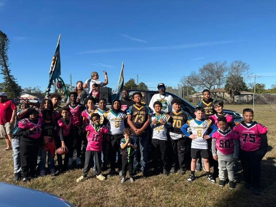 Group of children and adults gathered outdoors, with some children wearing football jerseys, standing in front of a vehicle with flags, under a clear blue sky.