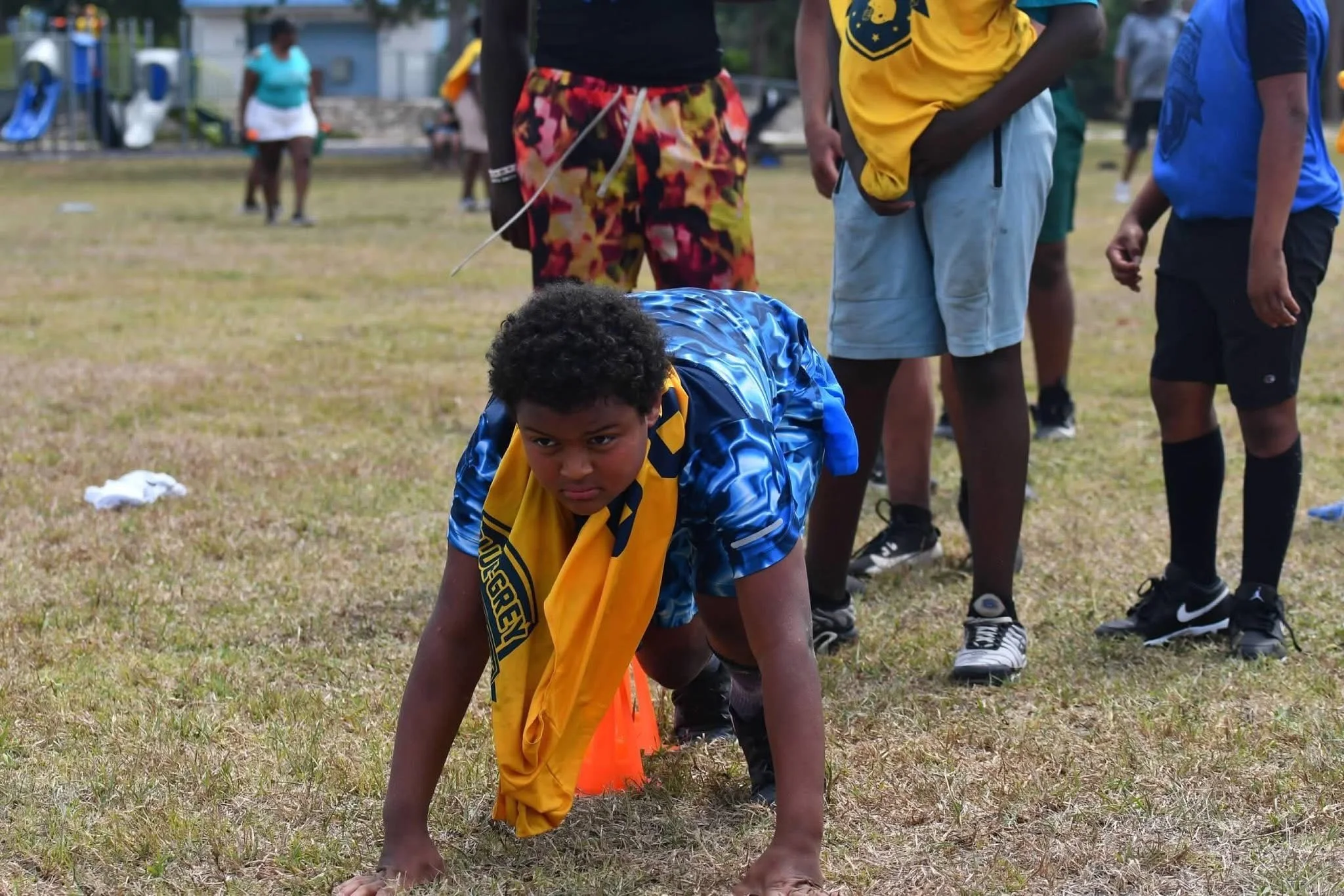 Young boy in blue athletic clothing crouches on the ground in a starting position at a race event, surrounded by other children and adults on a grassy field.
