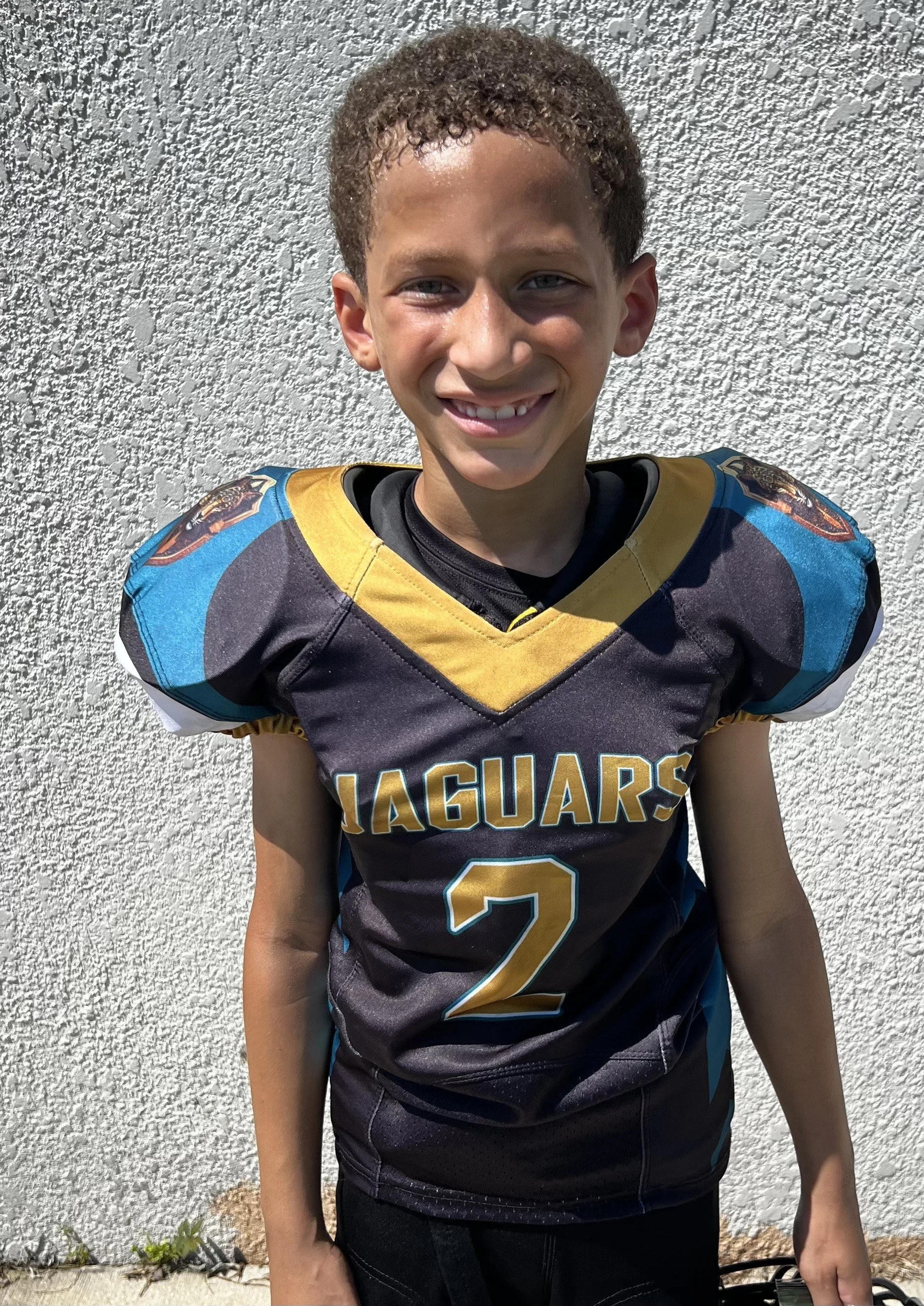 Young boy wearing a black and gold football jersey with 'Jaguars' and the number 2, standing against a white textured wall, smiling.