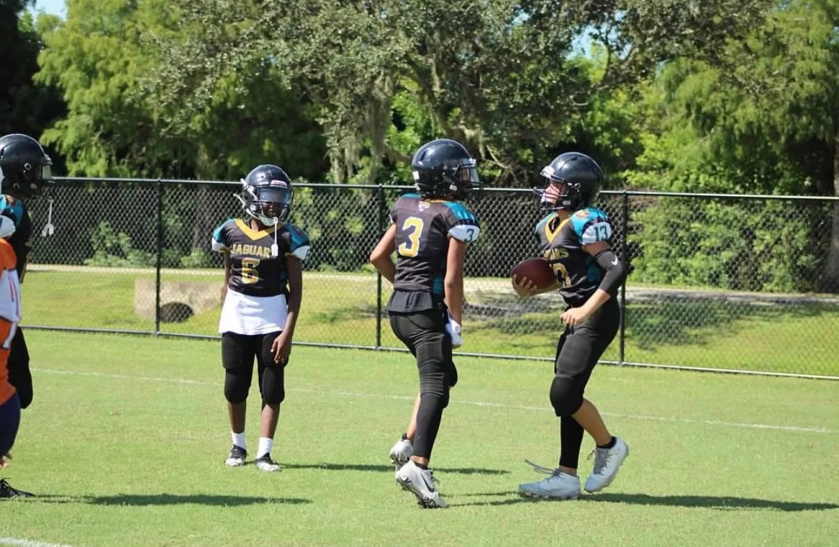 Youth football players on a field during practice or game, wearing helmets and jerseys with numbers, with a chain-link fence and trees in the background.