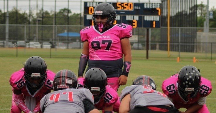 Football players in pink and gray uniforms preparing for a play on the field with a scoreboard in the background.