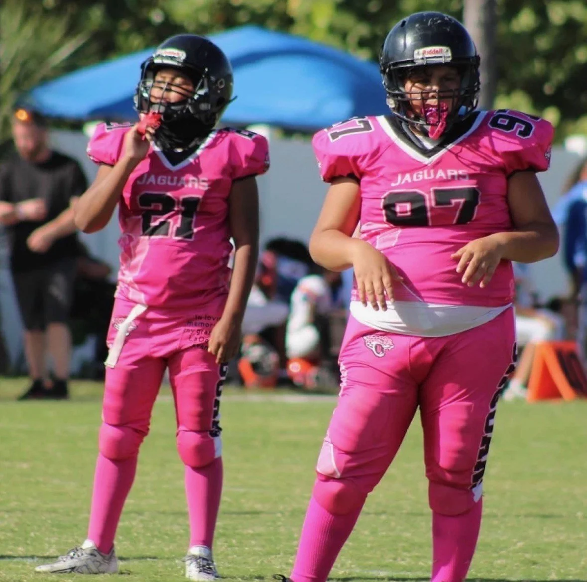 Two young female football players in pink jerseys with black helmets on a grassy field, celebrating or preparing for a play.