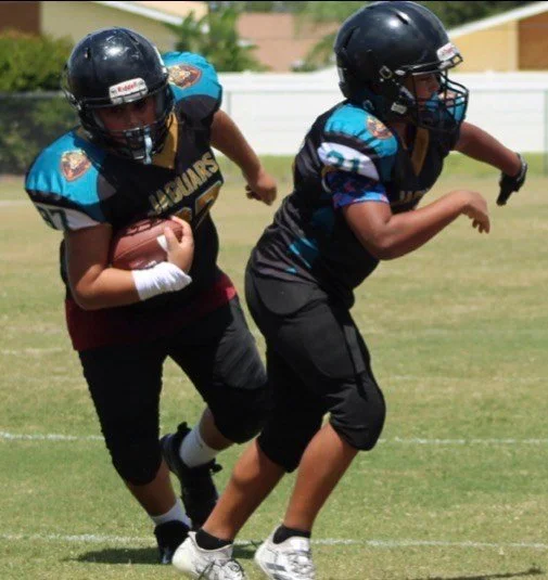 Two young boys playing youth football on a grassy field, both wearing helmets and team uniforms, one holding a football and running while the other tries to stop him.