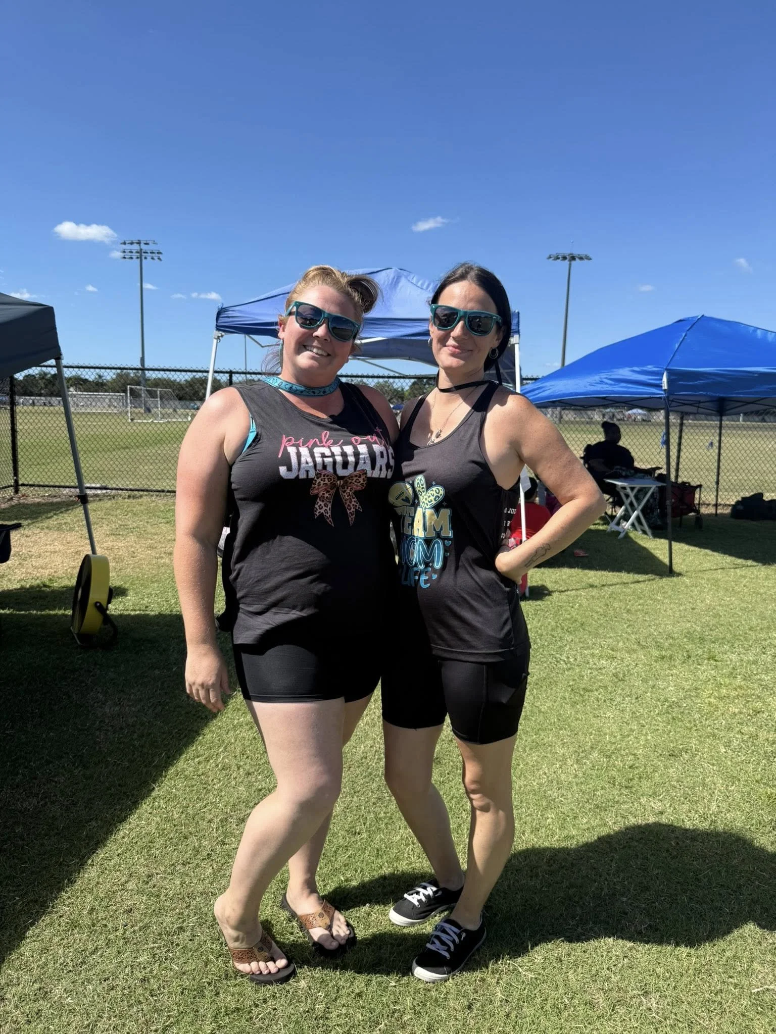 Two women wearing sunglasses and athletic clothing standing on grass at an outdoor sports field, with tents and a chain-link fence in the background under a clear blue sky.