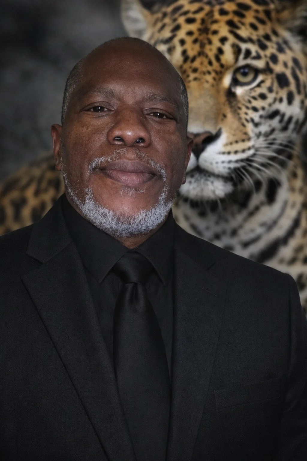 A man with gray hair and beard wearing a black suit and tie, standing in front of a large image of a leopard.