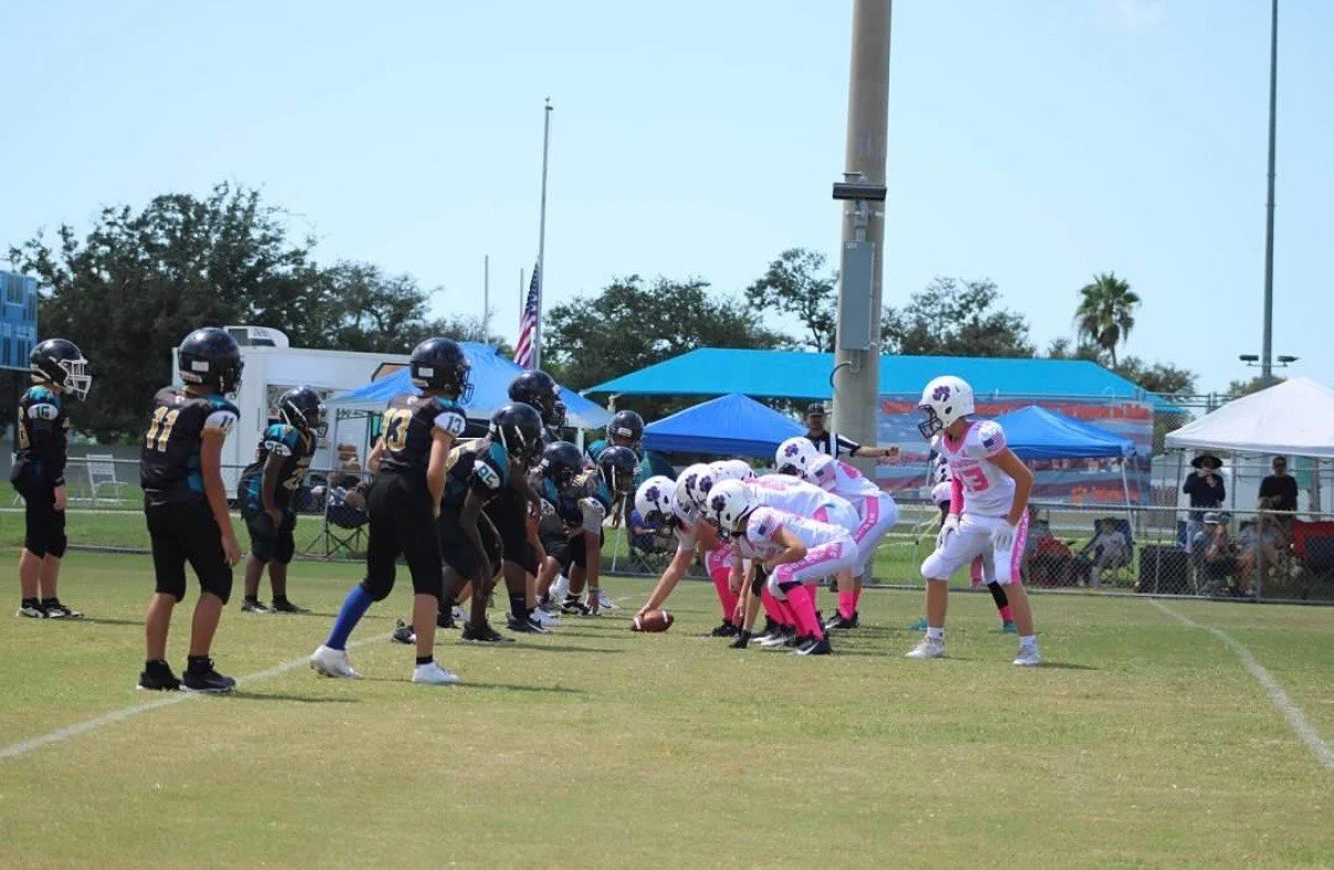 Youth football game on a grassy field with players in black and white uniforms, some stretching and preparing for the play, with blue tents and a fence in the background.