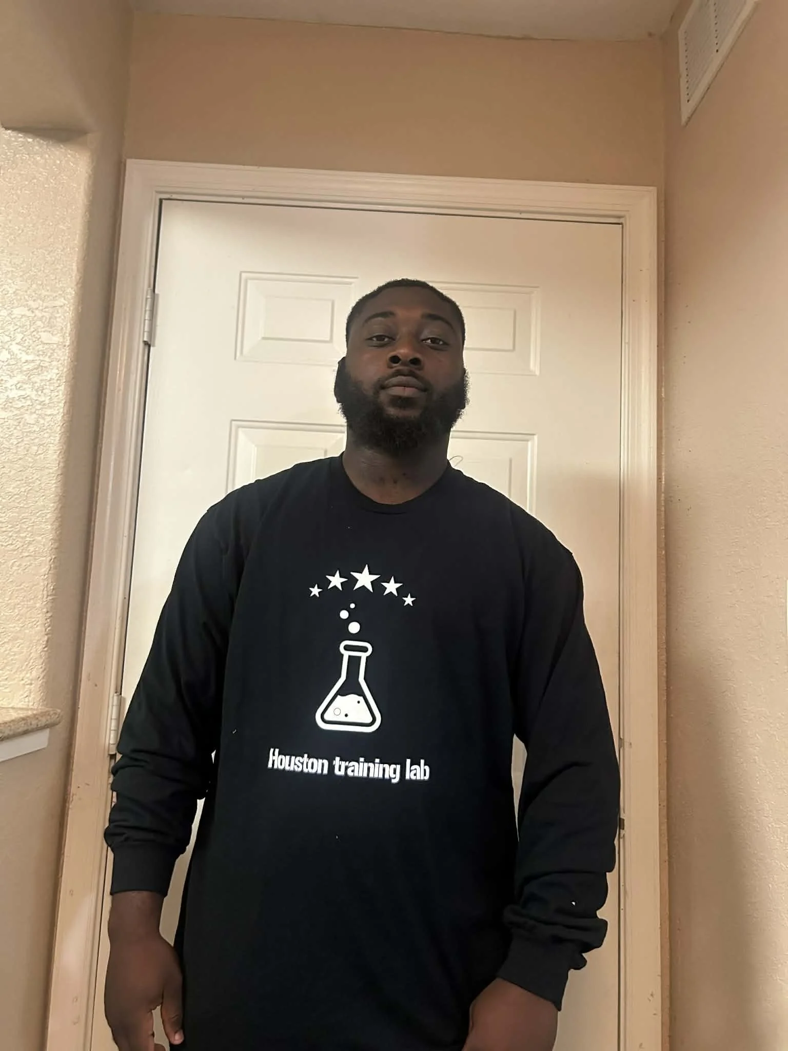 A man standing in front of a white door, wearing a black long-sleeve shirt with a science lab graphic and the words 'Houston training lab' on it.
